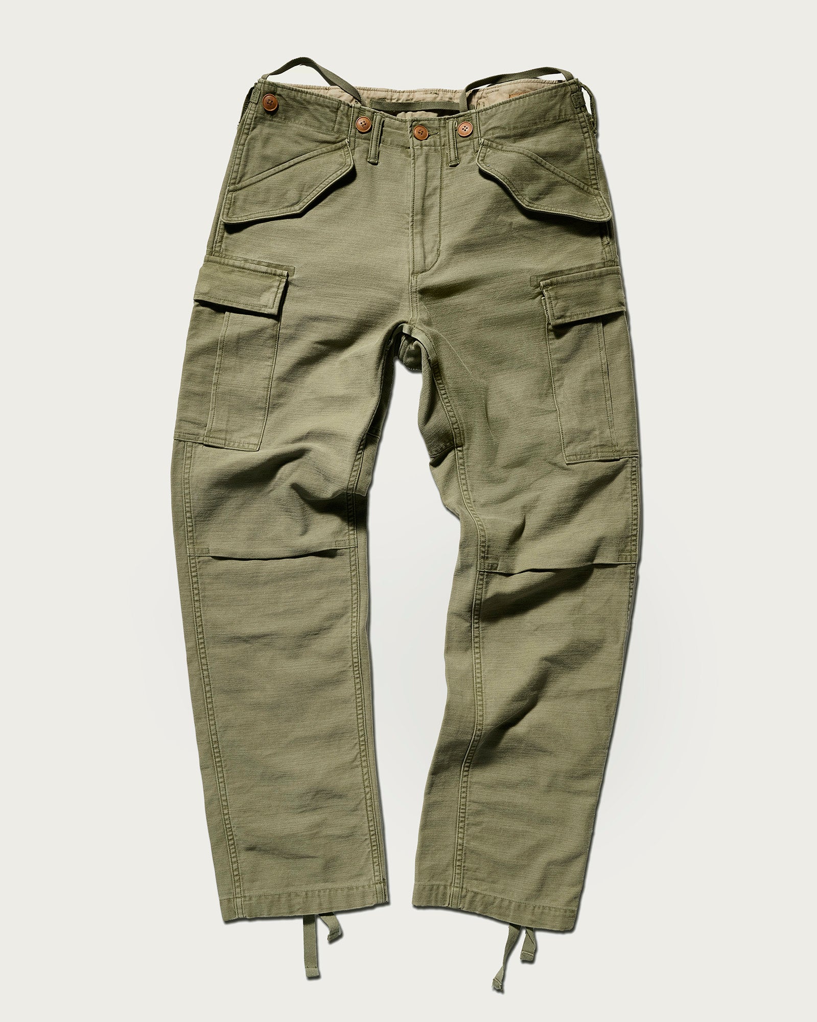 The Brando Cargo in Olive Drab by T7C, crafted from cotton-back satin with a sulphur-dyed finish, features multiple pockets, button and zip closure, adjustable waist tabs, and cuff drawstrings—shown laid flat on a light background.