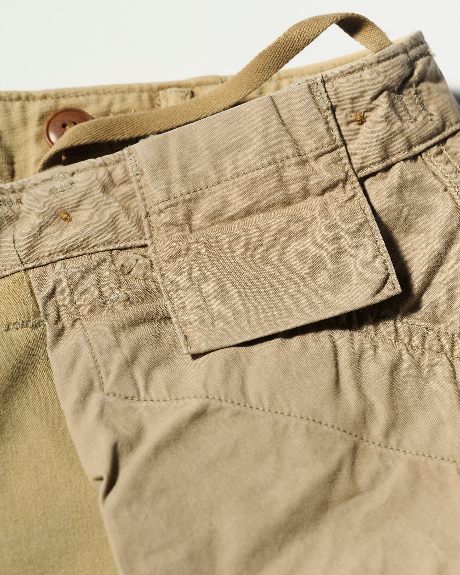 Close-up of The Brando Cargo in Sand by T7C, crafted from cotton-back satin. Features a button and flap pocket detail, visible stitching, textured fabric, a small loop, and part of another pant shown in the background.