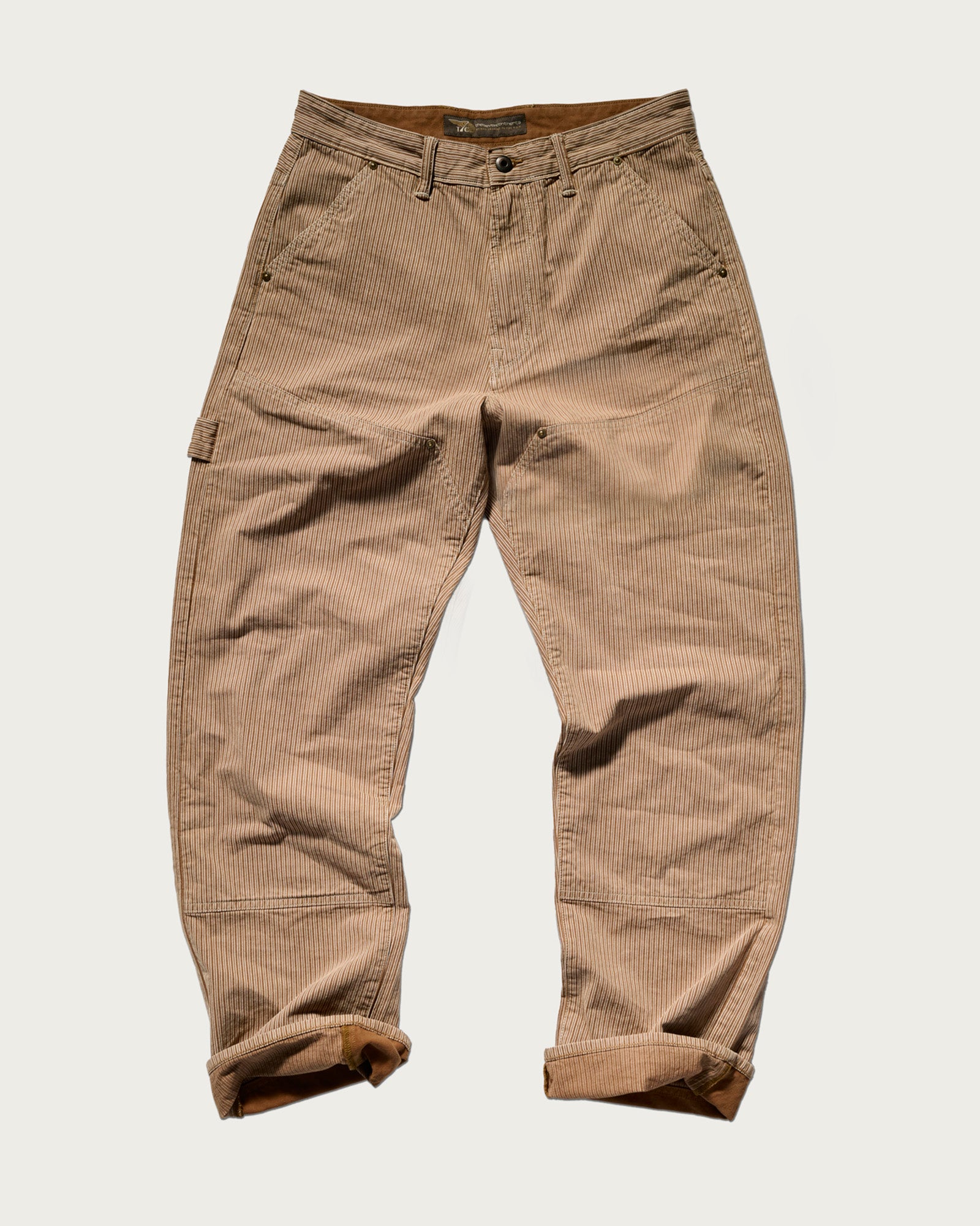 The T7C Fred Pant in Camel Stripe, shown flat on a white background, features front and side pockets, reinforced knees, a button-zipper closure, and rolled cuffs. The relaxed-fit carpenter style is done in tan and beige hickory railroad stripes.