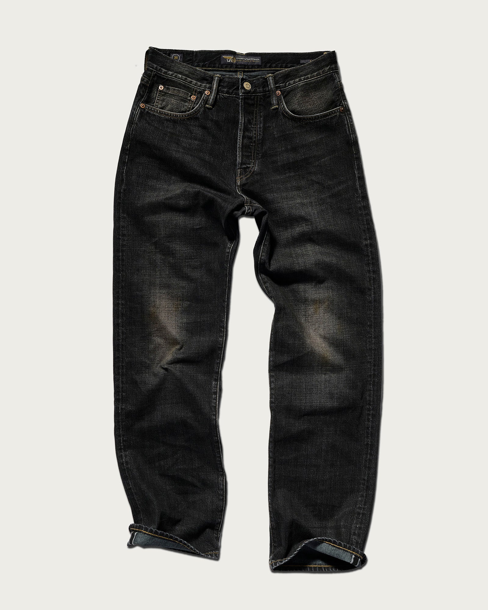 The Jack Jean In Kobe Black by T7C: a relaxed-fit, vintage-wash black jean with subtle thigh fading and rolled cuffs, shown on a plain light background.