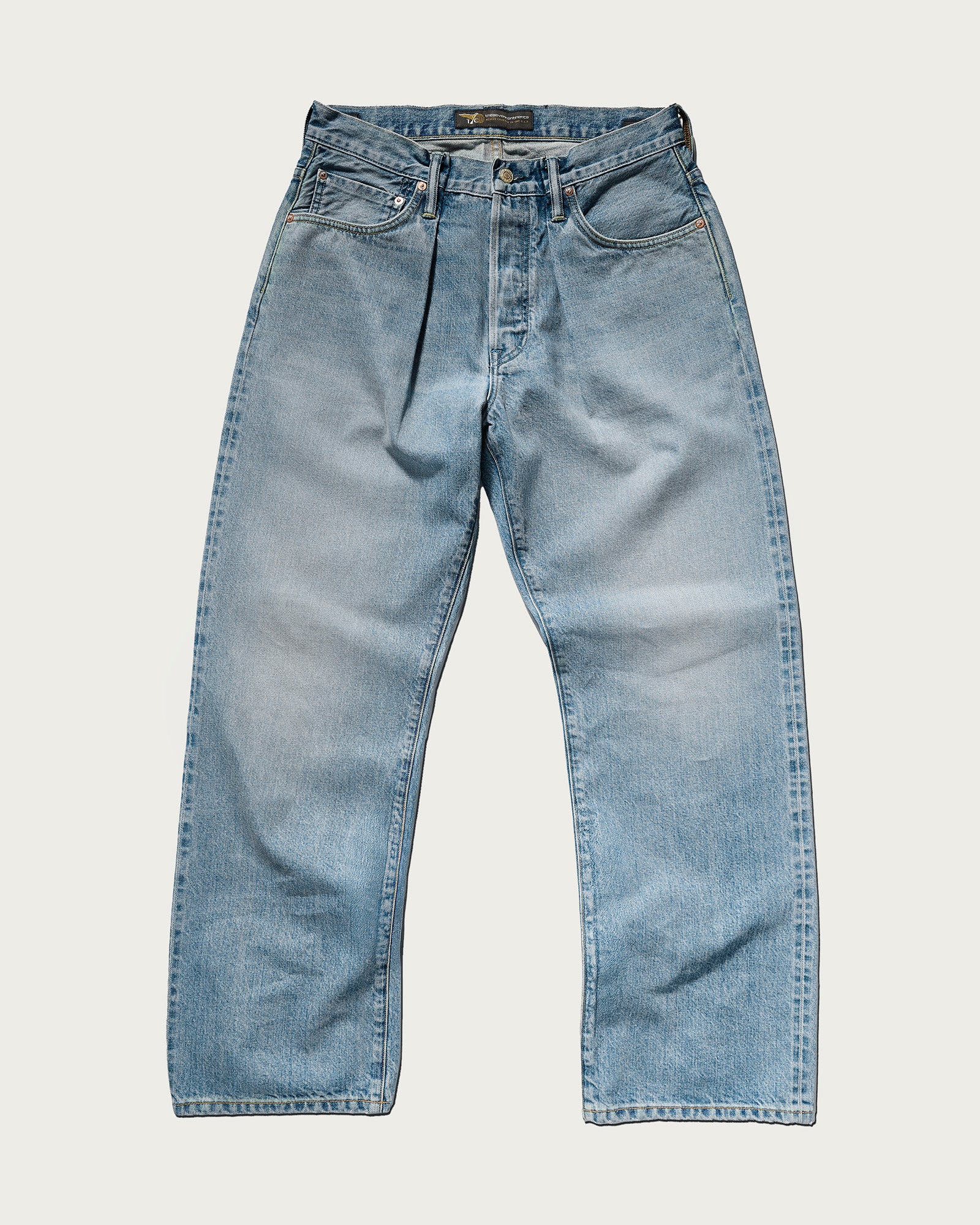 The Cinch Jean in Mykonos by T7C: light blue, straight-leg selvedge denim jeans with a button fly, front and back pockets, and faded detailing, shown laid flat on a white background.