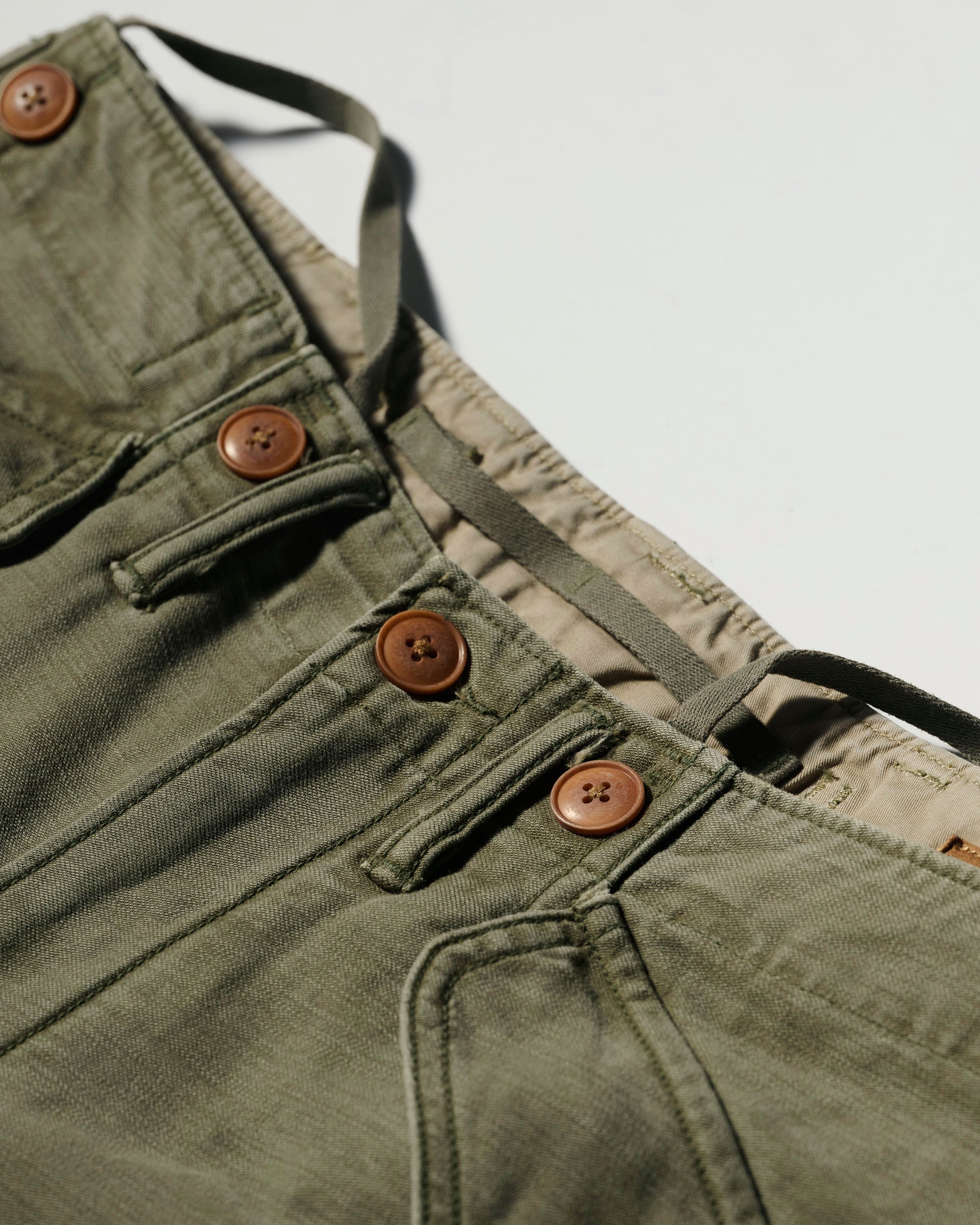 Close-up of The Brando Cargo in Olive Drab by T7C, featuring cotton-back satin fabric, brown buttons, front pockets, and an adjustable drawstring waistband against a light background.