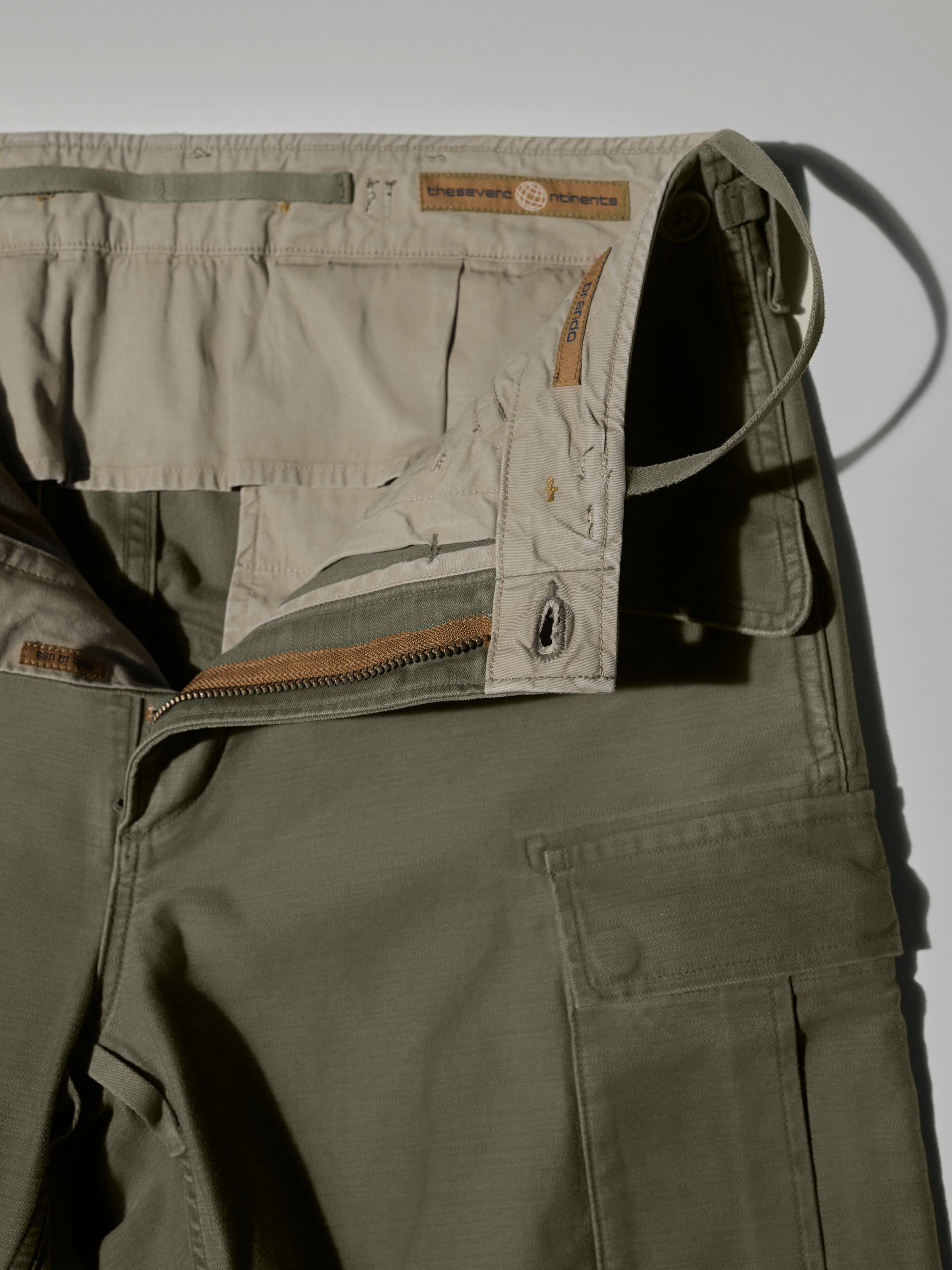 Close-up of The Brando Cargo in Olive Drab by T7C, crafted in cotton-back satin with a partially unzipped fly, beige inner waistband, and visible Reserved Nature label. Features sulphur dyeing, side pocket, and utilitarian button details.