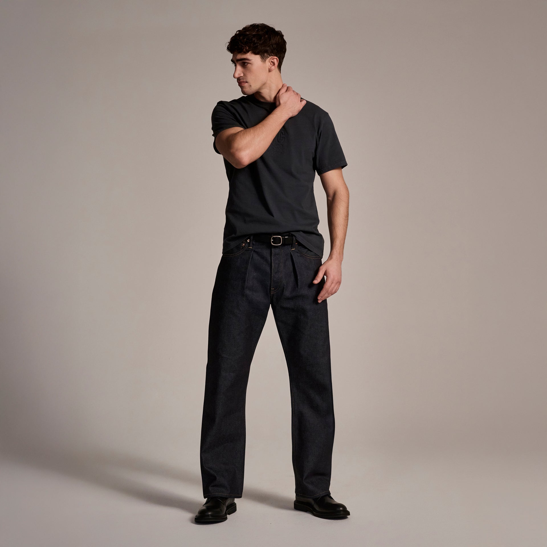 A young man stands in a neutral studio, looking left and touching his neck, wearing a black t-shirt, black shoes, and T7C The Seven Continents The Cinch Jean in Yokohama Raw.