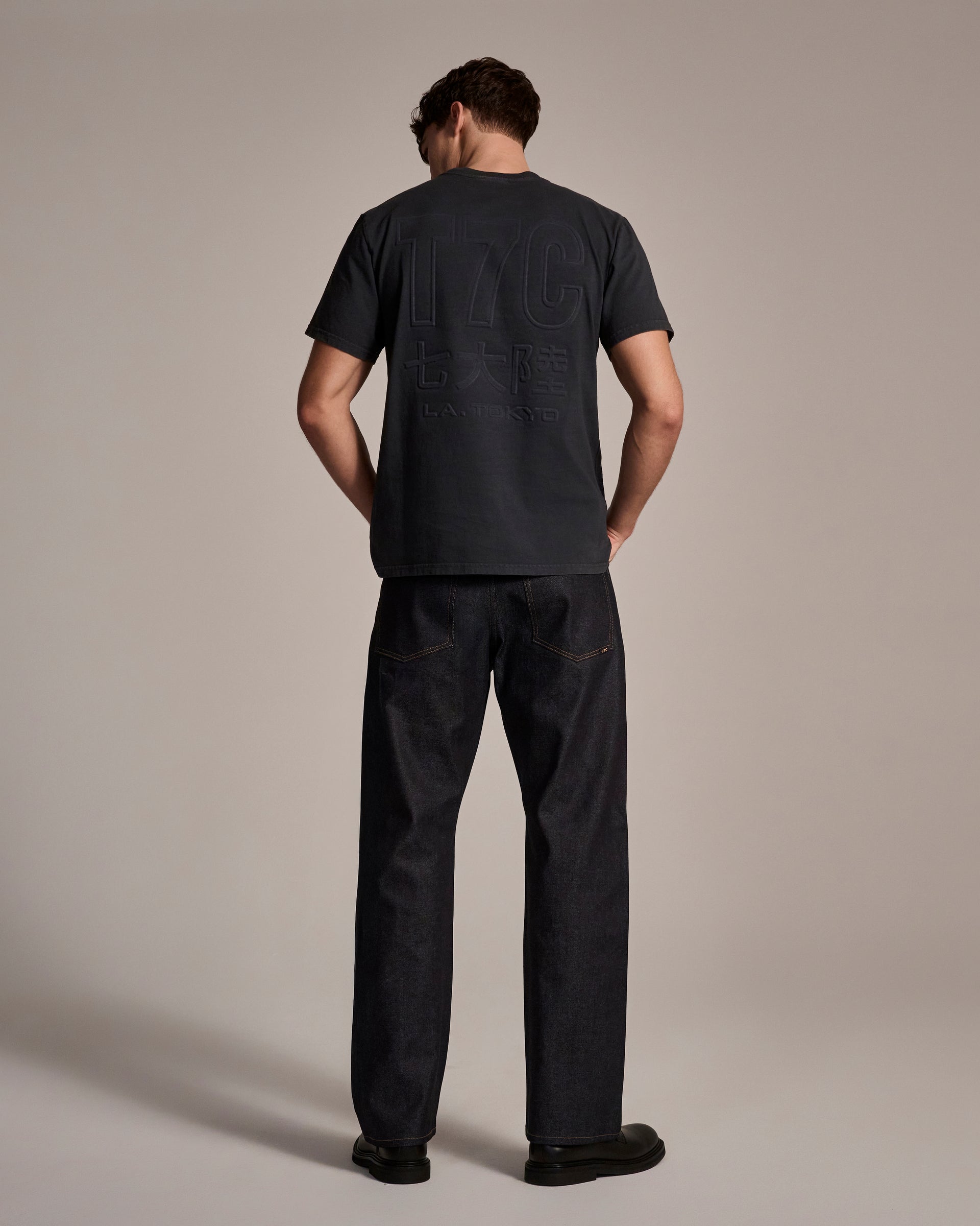 A man with short brown hair wears T7C The Seven Continents Cinch Jean in Yokohama Raw and a dark embossed short-sleeve tee. He faces away, posed against a plain neutral background, in black shoes and oversized indigo-dyed denim jeans.