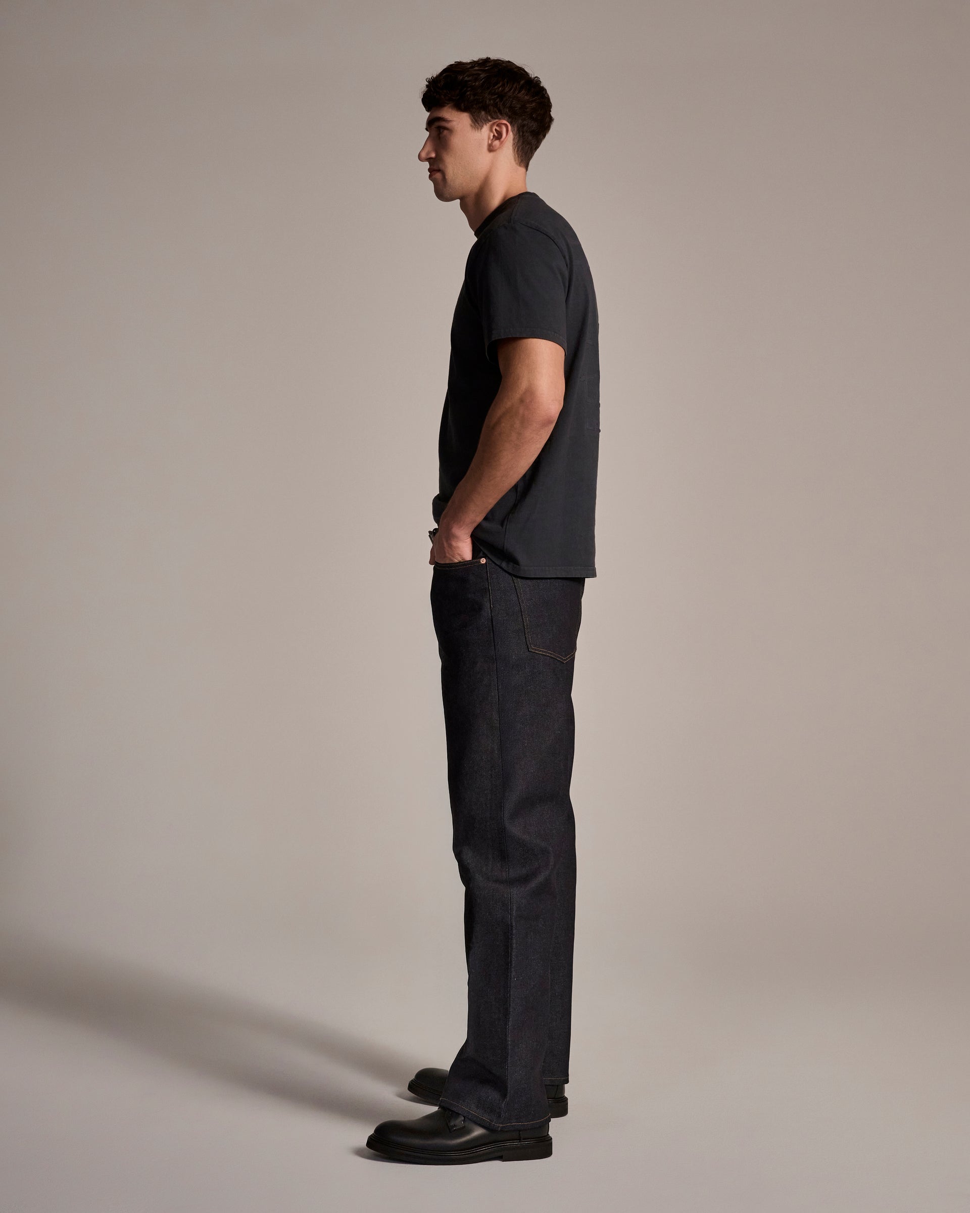 A man stands in profile against a plain background, wearing T7C The Seven Continents’ The Cinch Jean in Yokohama Raw, a black T-shirt, and black shoes with his hands in his pockets, looking straight ahead.