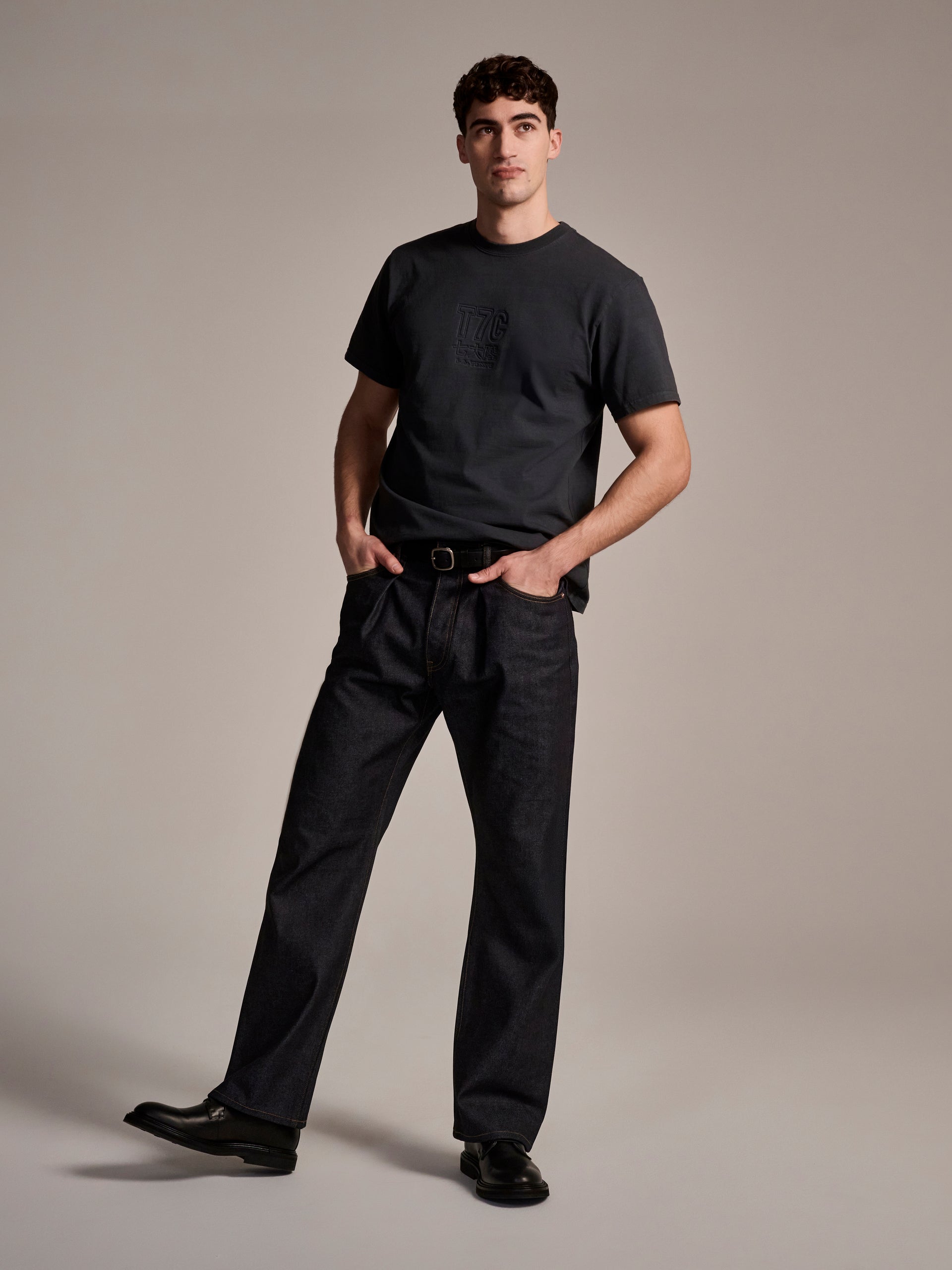 A man stands confidently against a neutral background, wearing T7C The Seven Continents The Cinch Jean in Yokohama Raw—an oversized fit Japanese selvedge denim—paired with a black t-shirt, black belt, and black shoes, hands in pockets.