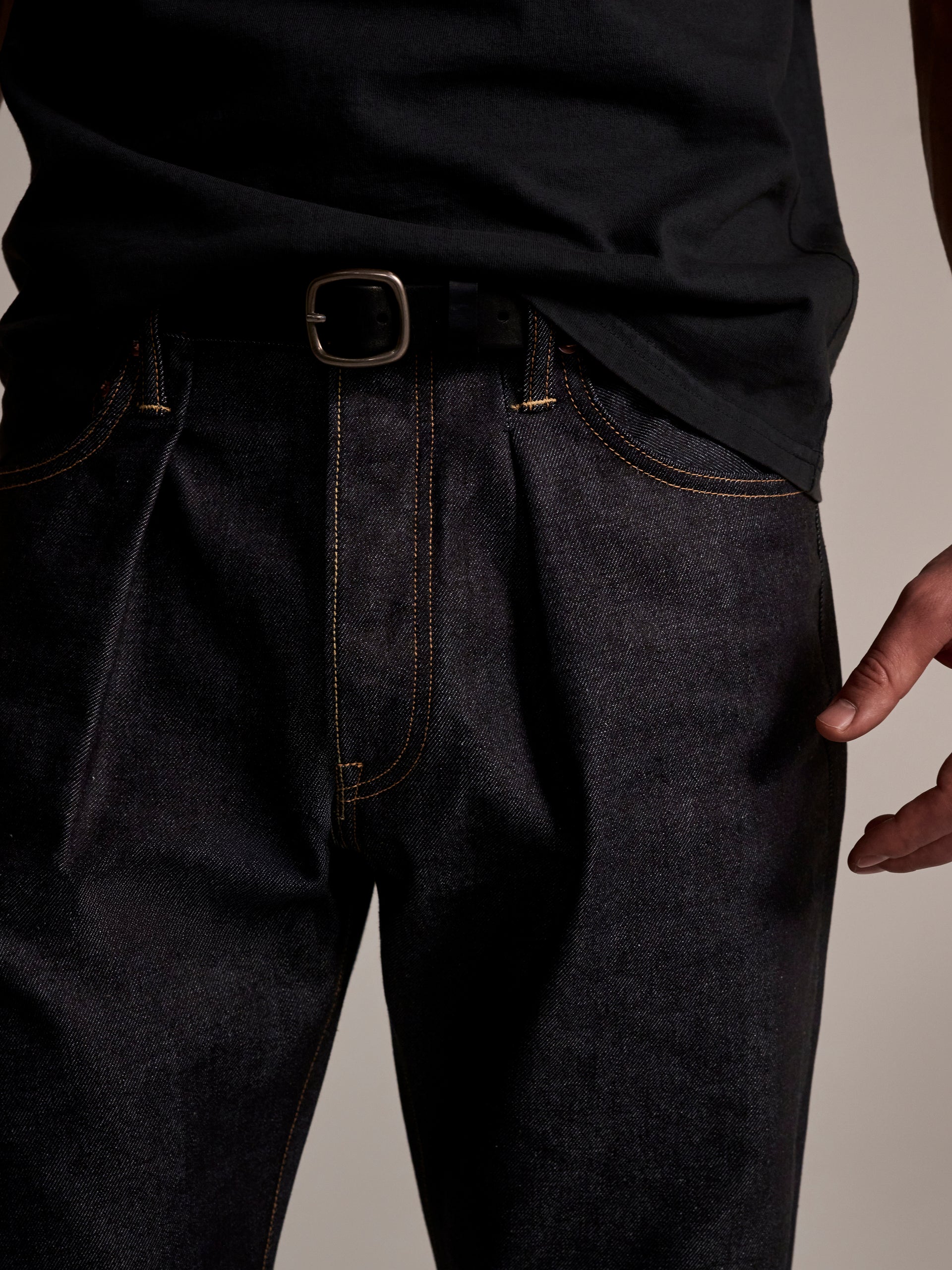 A person is shown from waist to mid-thigh wearing a black t-shirt and T7C The Seven Continents The Cinch Jean in Yokohama Raw, styled with yellow stitching and a black belt with a silver buckle. Their right arm rests relaxed at their side.