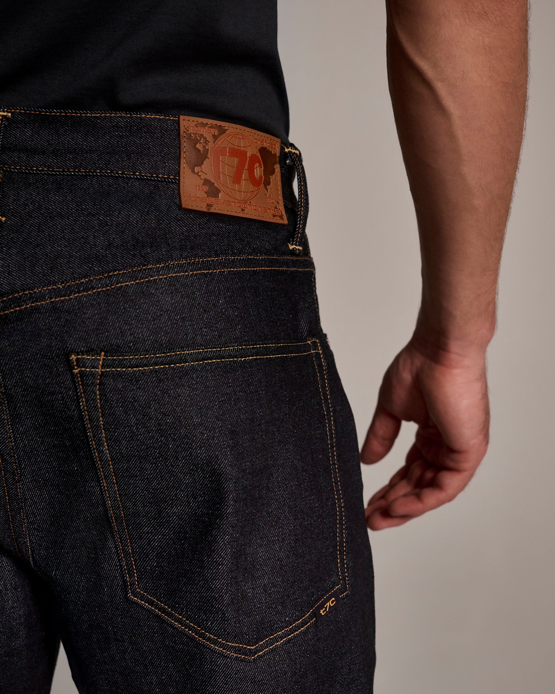 A close-up of a person wearing T7C The Seven Continents’ The Cinch Jean in Yokohama Raw, featuring indigo-dyed denim, bold stitching, and a brown leather waistband patch, with a hand resting by their side.