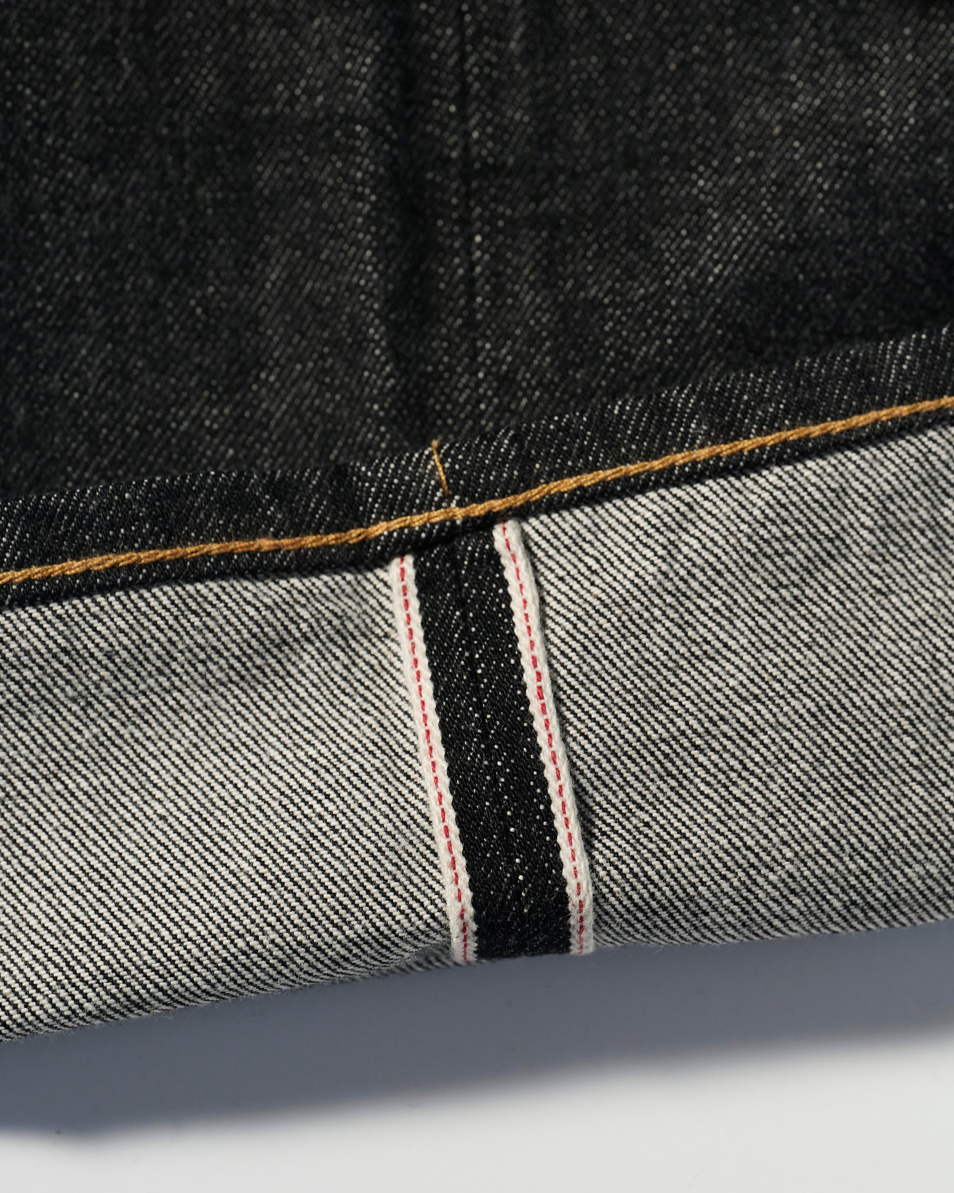A close-up of The Charlie Jean in Fuji Black by T7C highlights the cuffed dark denim—crafted from organic Turkish cotton—with white selvedge, red stitching, and yellow topstitching on the edge.