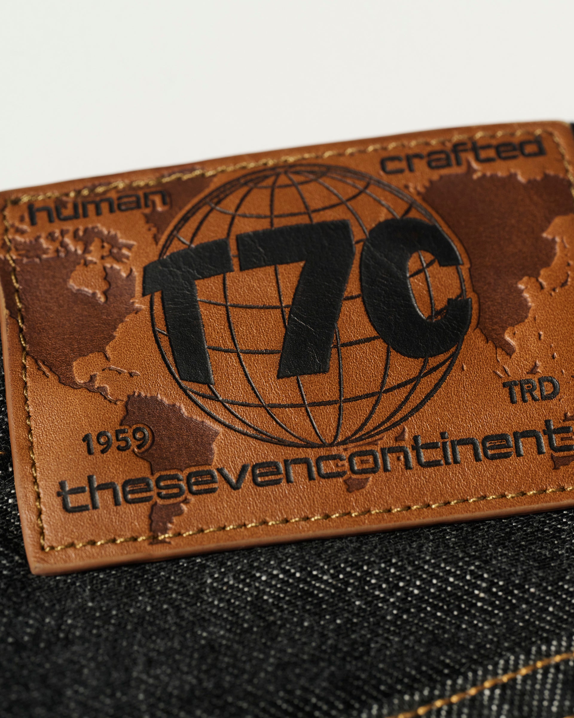 Close-up of a brown leather patch on The Charlie Jean in Fuji Black by T7C, embossed with a world map, globe, bold T7C and thesevencontinents text, plus details like human crafted and 1959—crafted from organic Turkish cotton.