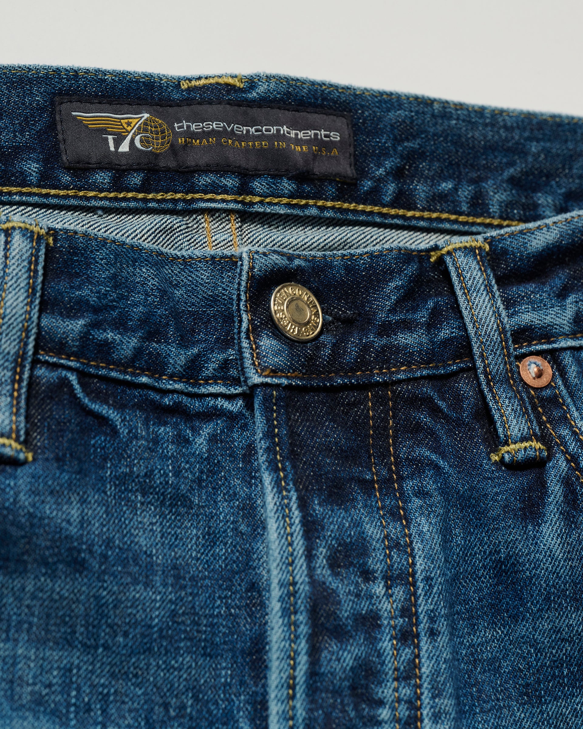 Close-up of The Charlie Jean in Fuji Medium by T7C shows blue denim, a metal button, front pockets, yellow stitching, and a black thesevencontinents label with added text and a small graphic.