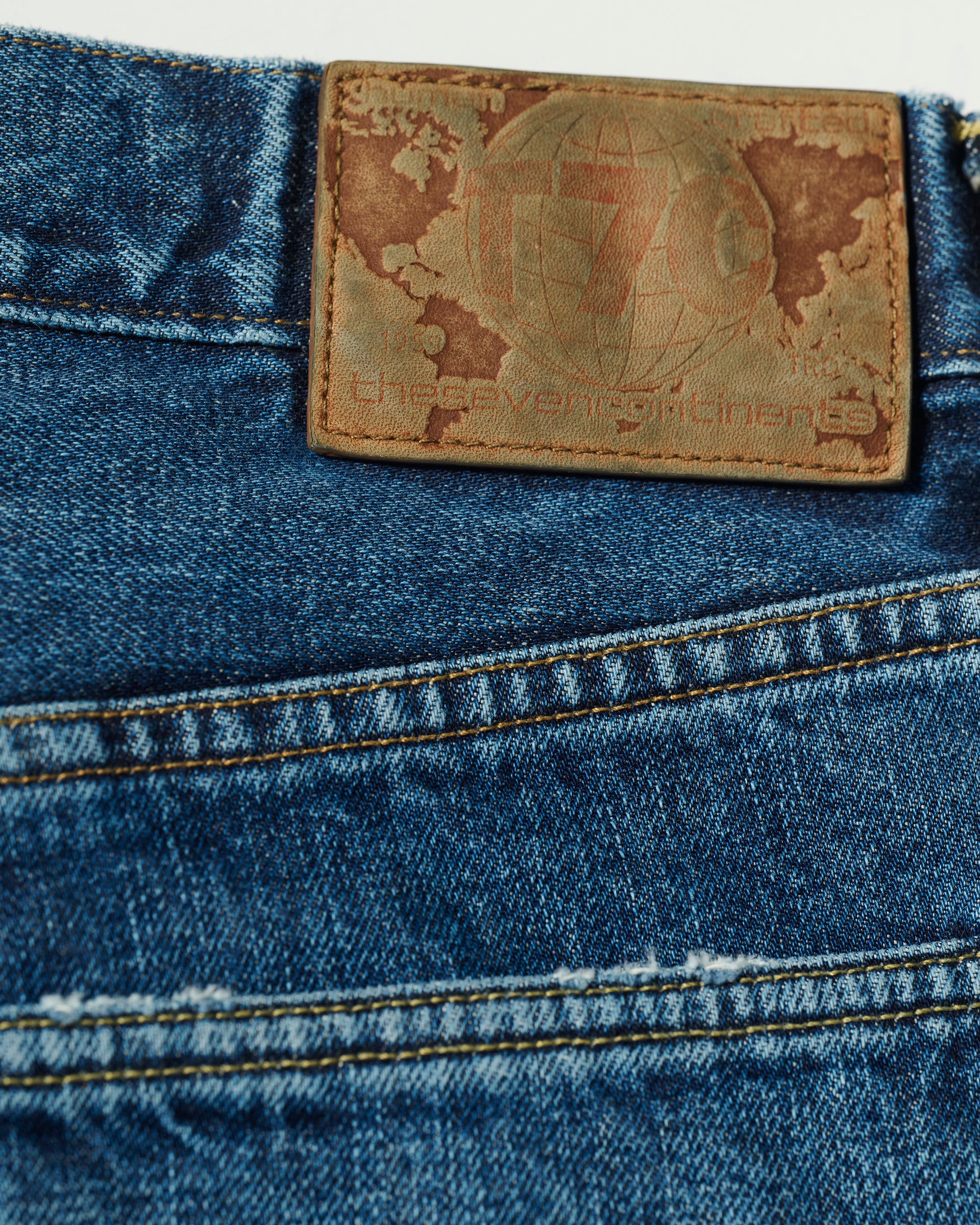 Close-up of The Charlie Jean in Fuji Medium by T7C—blue slimmest fit jeans with a brown leather patch on the waistband featuring the brands faded design and text. Visible stitching and texture highlight their tailored style.