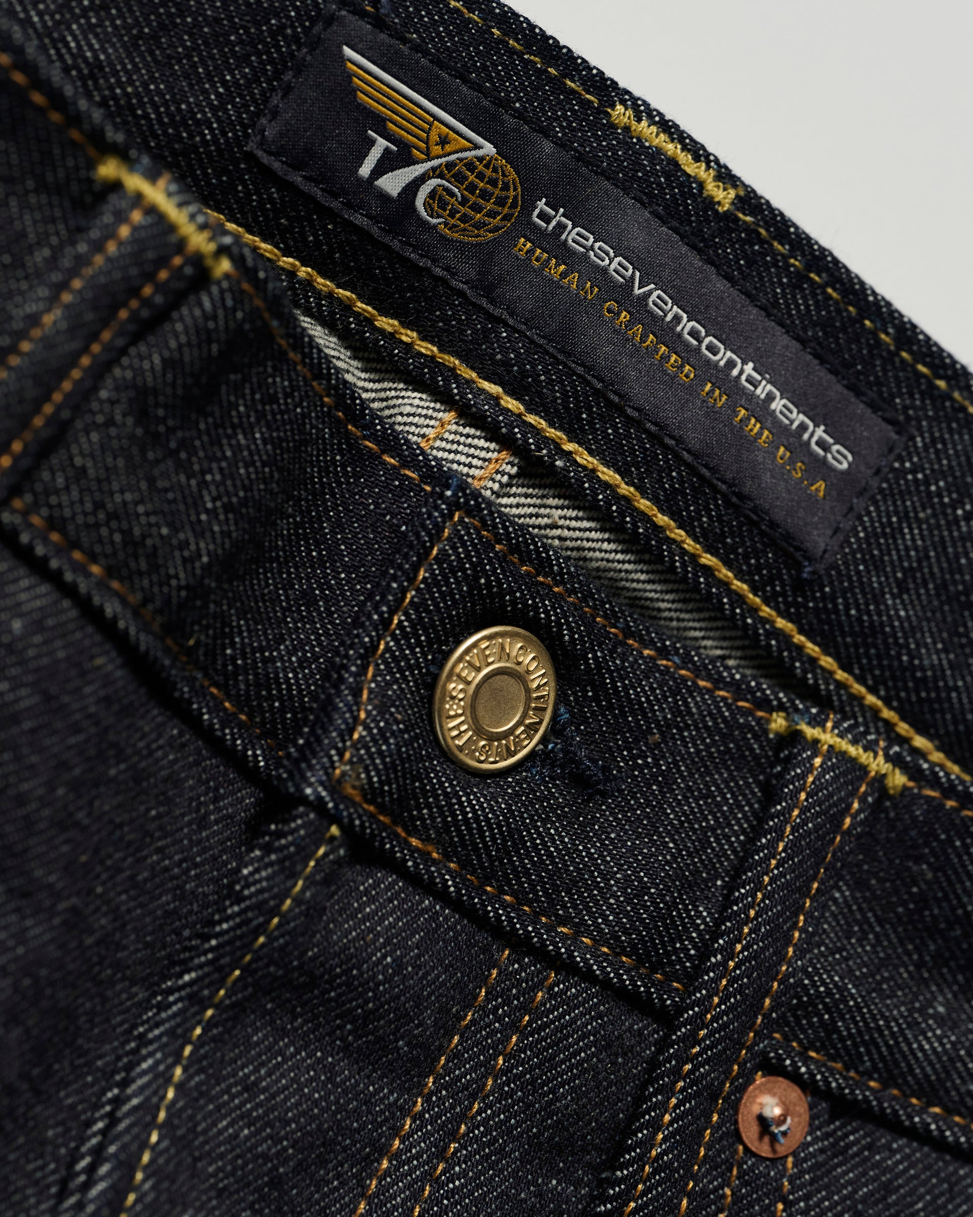 Close-up of The Charlie in Yokohama Raw by T7C The Seven Continents: indigo rope-dyed slim fit jeans in dark blue with a brass button, orange stitching, and a black thesevencontinents HUMAN CRAFTED IN THE U.S.A. label near the waistband.