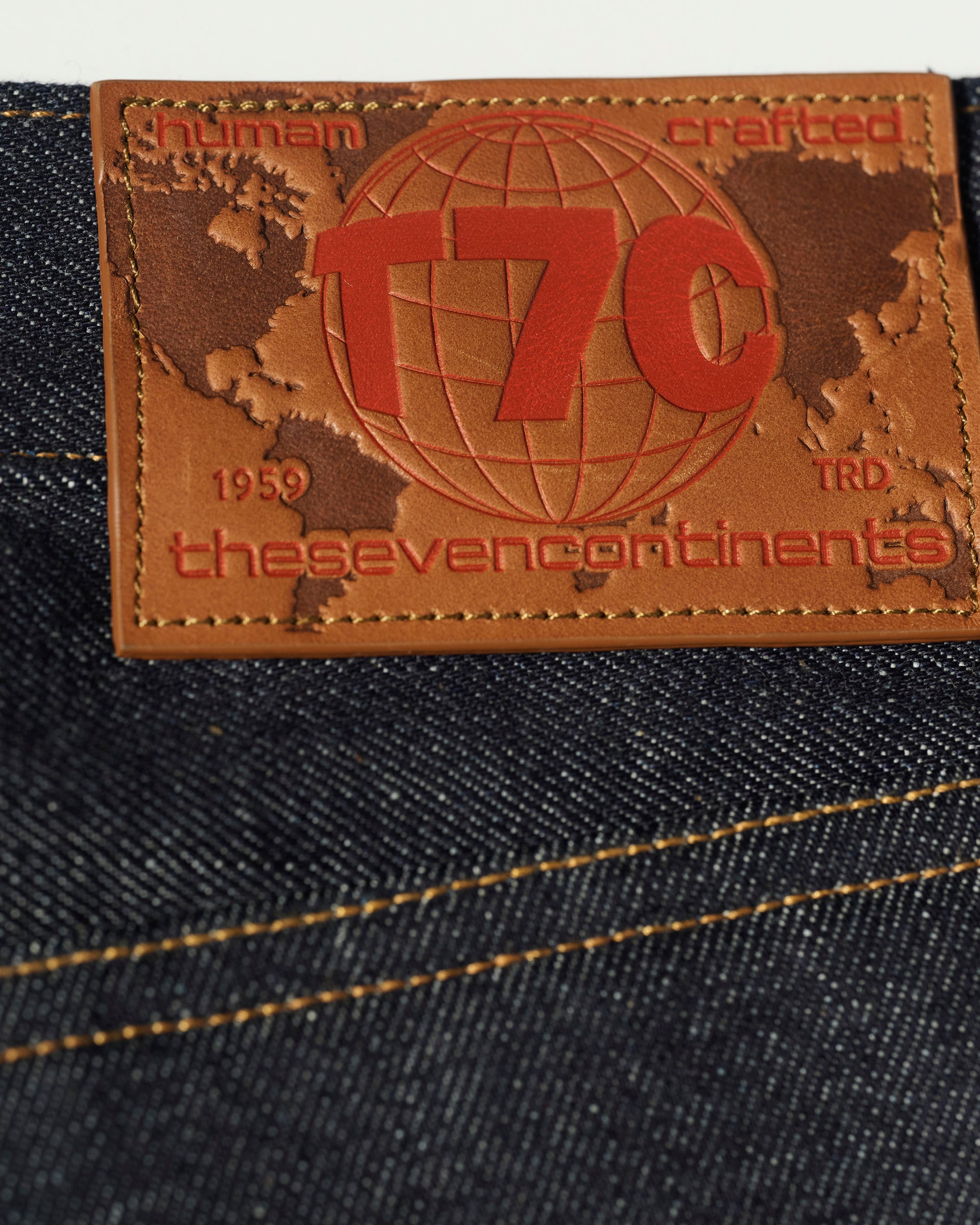 Close-up of a leather patch embossed with T7C, human crafted, 1959, thesevencontinents, and TRD on indigo rope-dyed Japanese selvedge denim fabric. Product: The Charlie in Yokohama Raw by T7C The Seven Continents.