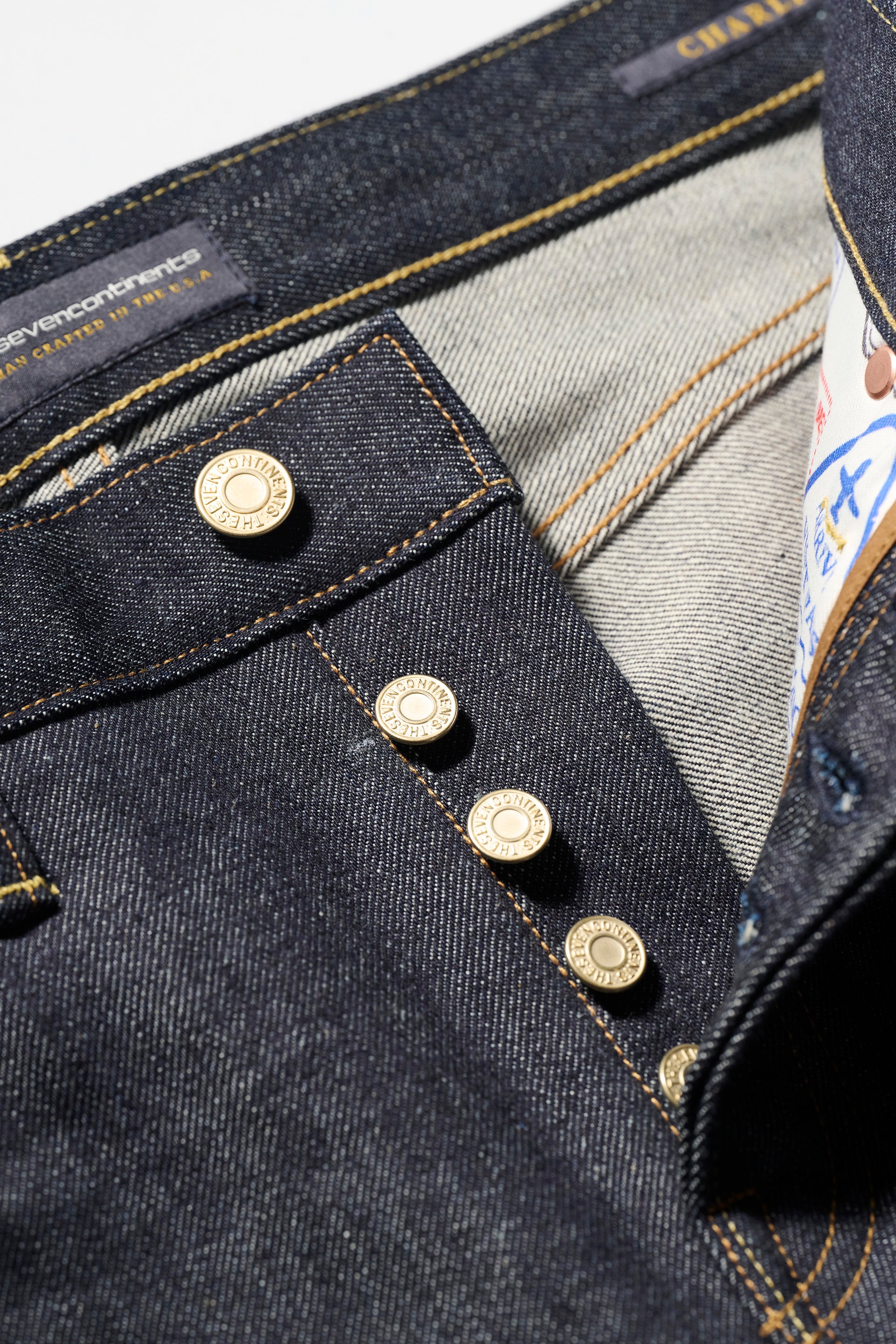 Close-up of The Charlie in Yokohama Raw by T7C The Seven Continents: slim fit jeans in dark indigo Japanese selvedge denim with four metallic fly buttons, yellow stitching, a light interior, and partial printed waistband label.