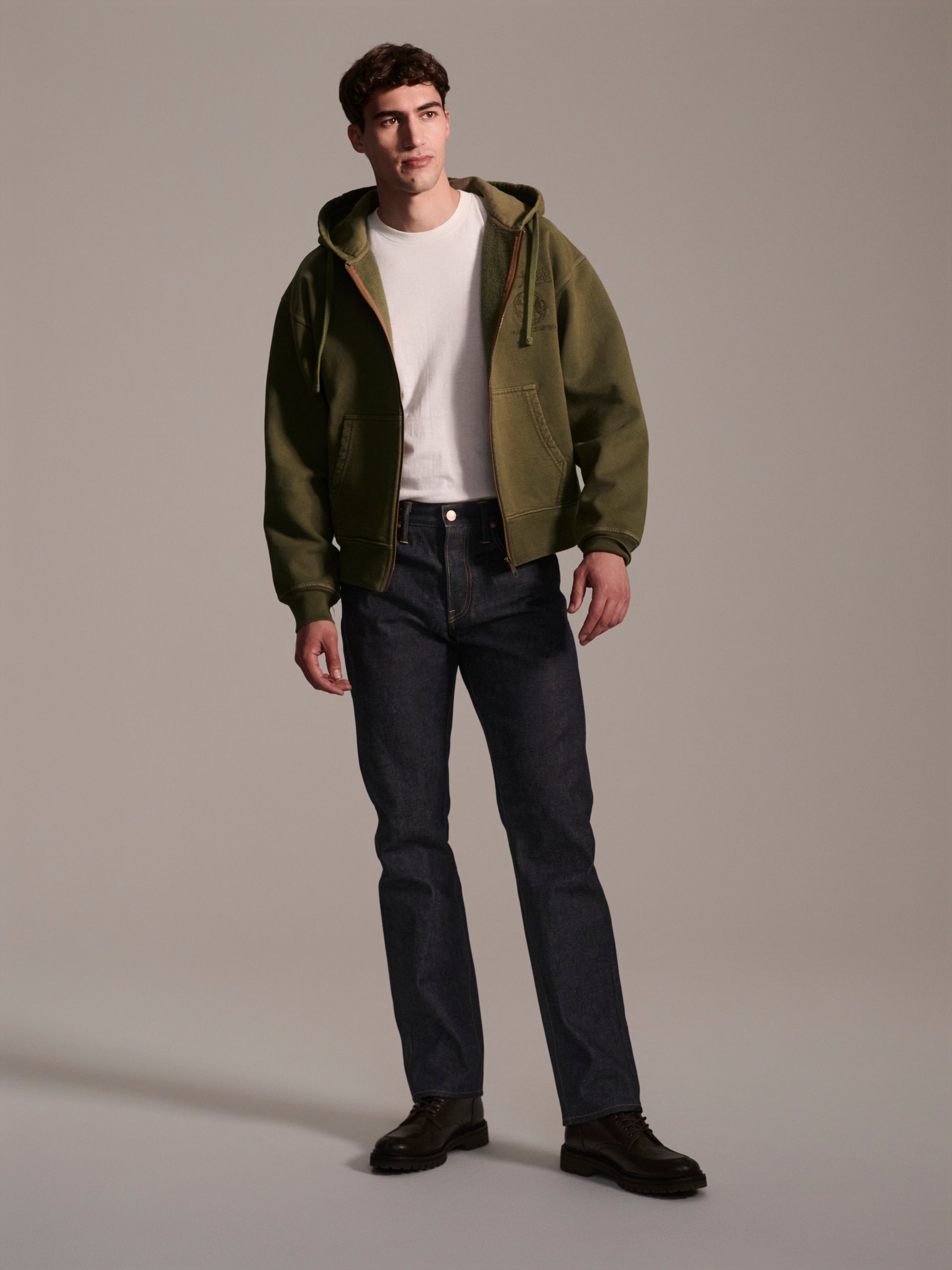 A man stands against a light gray background wearing a green zip-up hoodie, white t-shirt, black shoes, and T7C The Seven Continents The Charlie in Yokohama Raw Japanese selvedge denim slim fit jeans in dark blue.