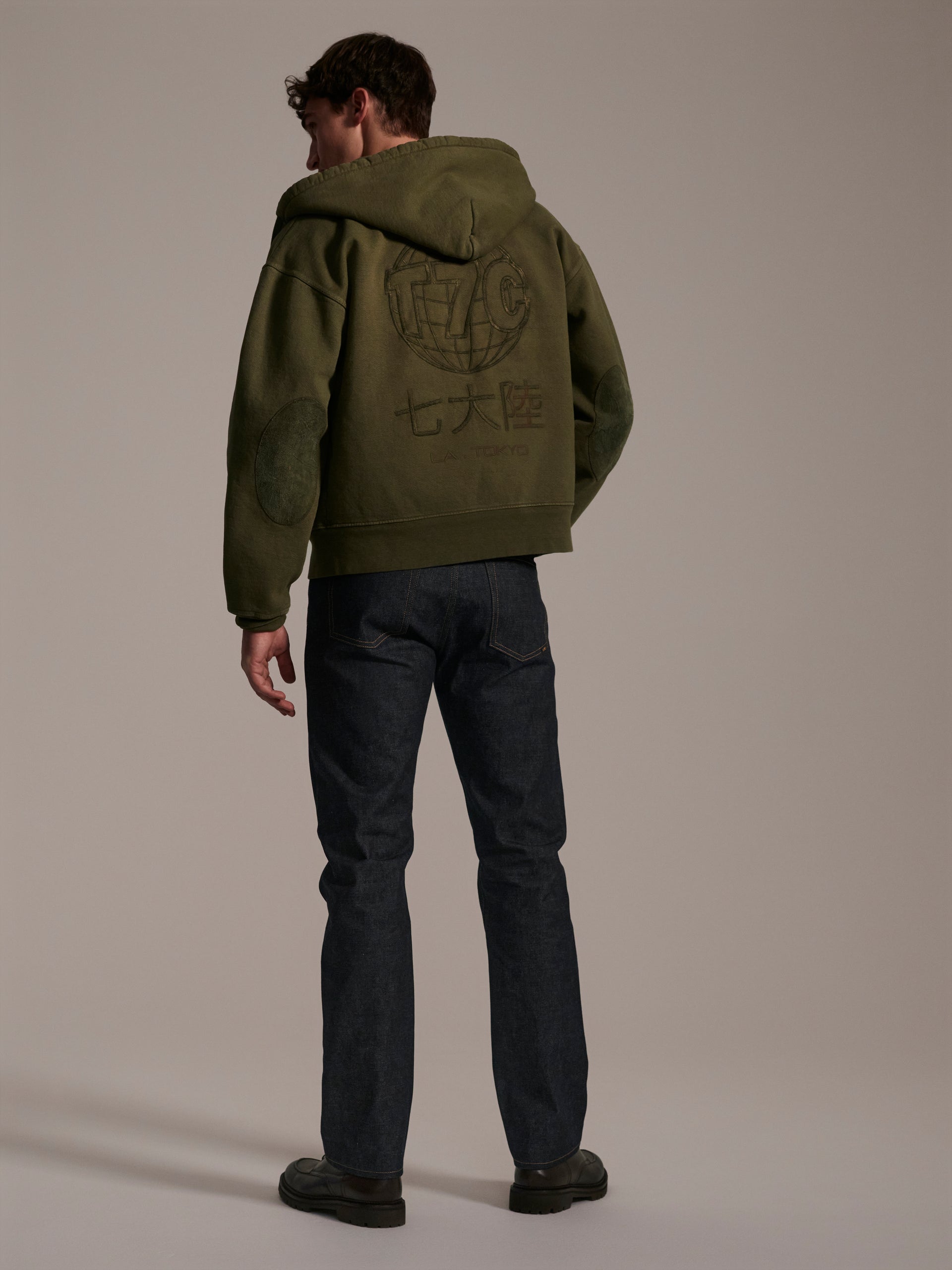 A person faces away in a green hooded jacket with graphic designs and Asian characters, paired with T7C The Seven Continents The Charlie in Yokohama Raw indigo rope-dyed Japanese selvedge denim slim fit jeans and dark shoes in a neutral studio.