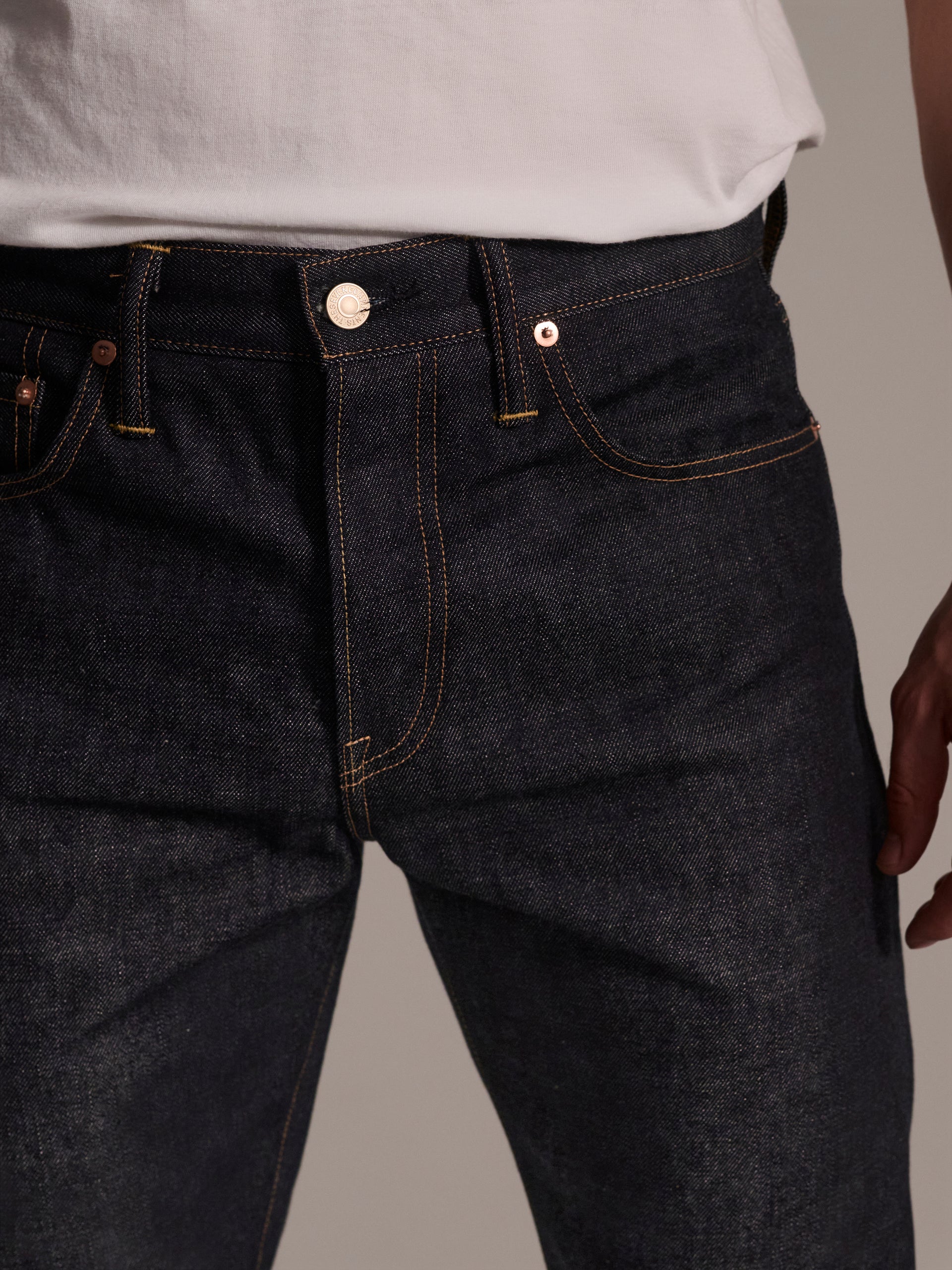 A person wearing a white shirt and T7C The Seven Continents The Charlie in Yokohama Raw indigo rope-dyed slim fit Japanese selvedge jeans with yellow stitching and classic copper rivets, shown waist to mid-thigh.