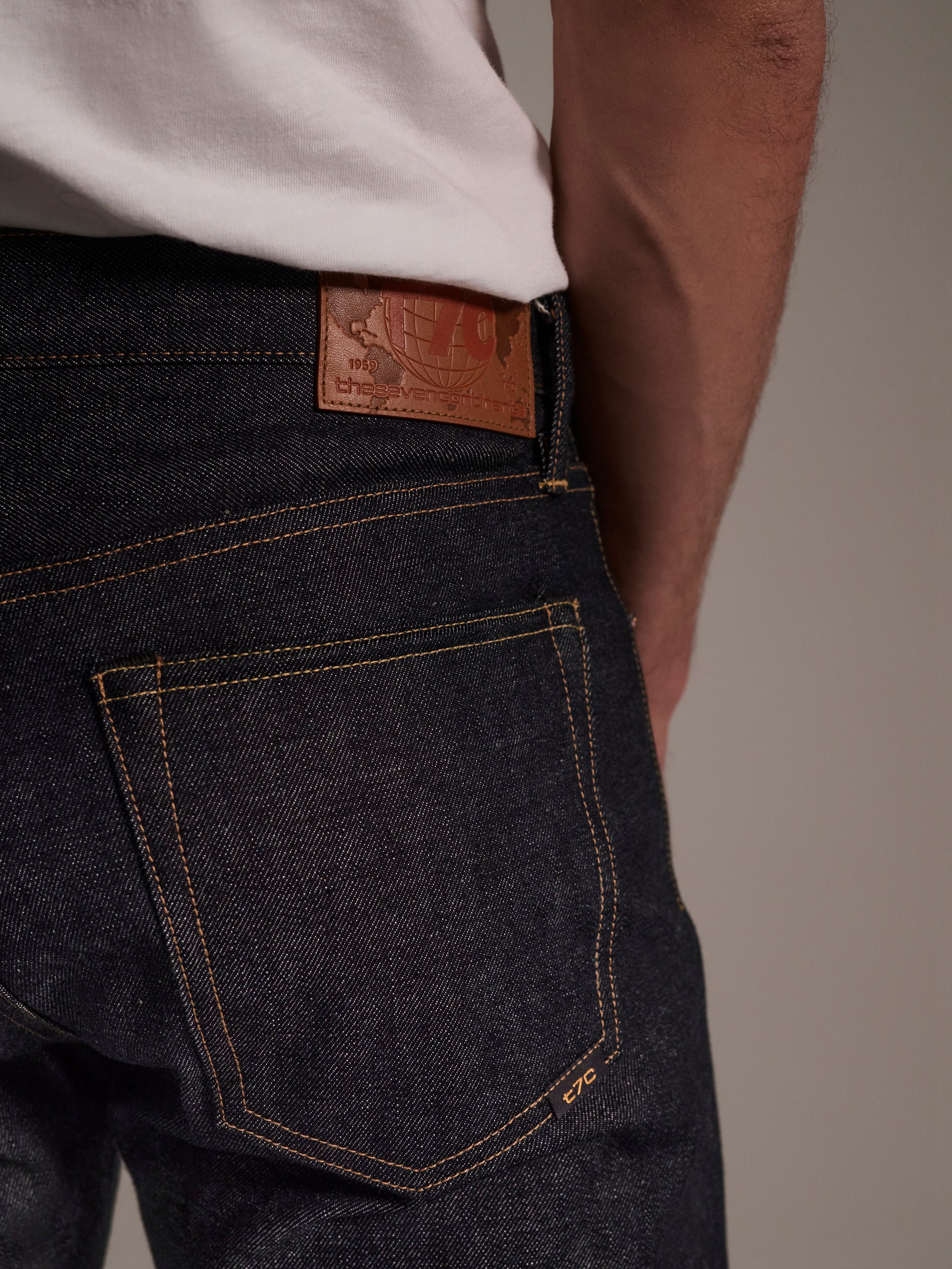 A close-up of a person wearing T7C The Seven Continents The Charlie in Yokohama Raw slim fit jeans, featuring indigo Japanese selvedge denim, yellow stitching, a brown leather patch on the waistband, and a white shirt tucked in.