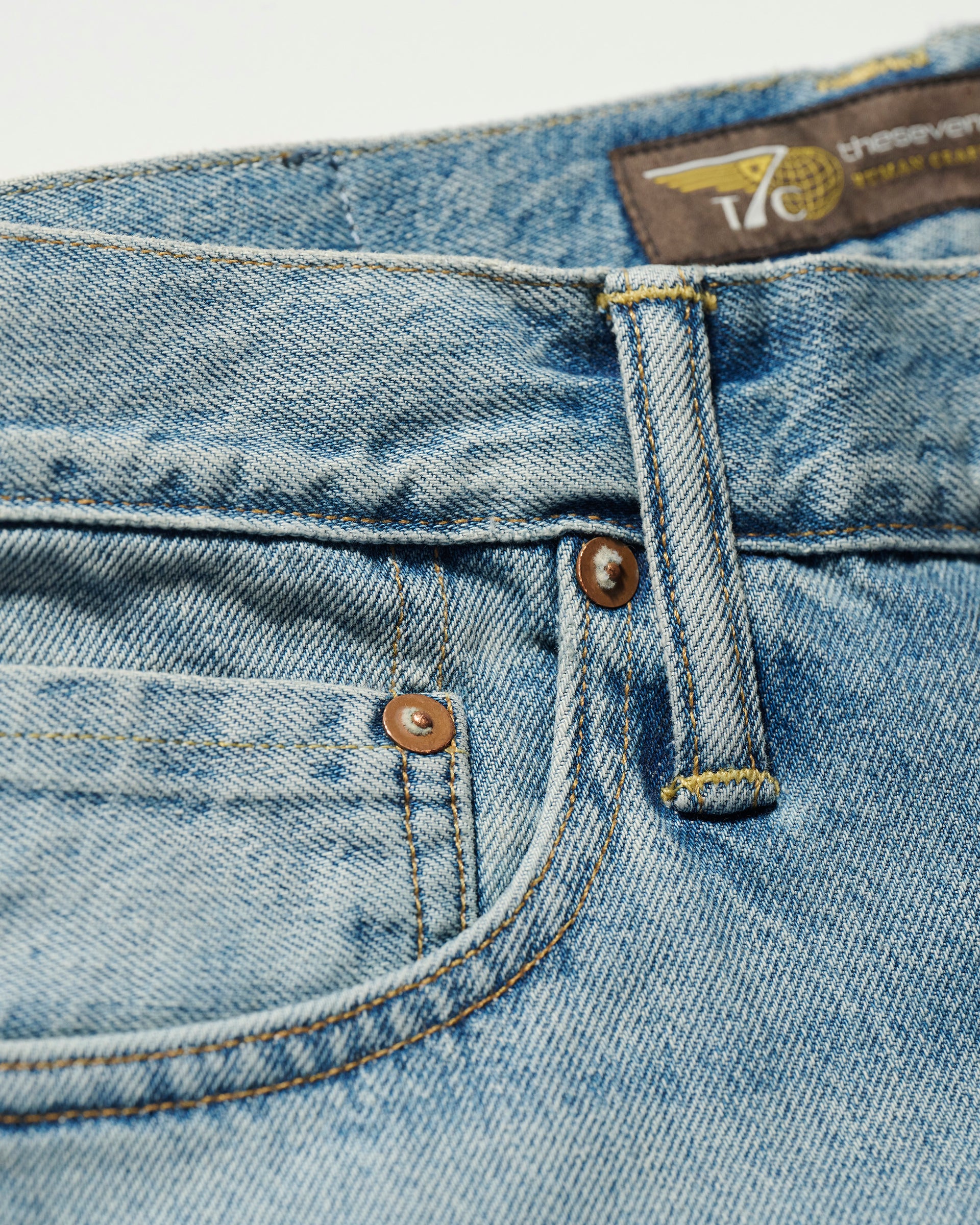 Close-up of the front pocket and waistband of T7C The Seven Continents The Charlie Jean in Zuma, light blue Japanese denim, showing stitching, rivets, belt loop, and part of a brown label with yellow text and a small graphic.