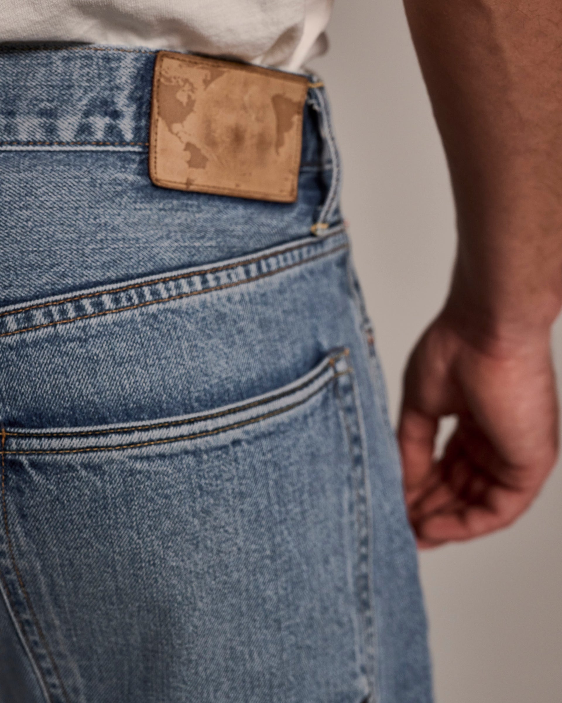 Close-up of a person wearing T7C’s The Cinch Jean in Mykonos—light blue selvedge denim with a white shirt, featuring the back pocket and a worn brown leather patch on the waistband. The person’s arm is partially visible at their side.