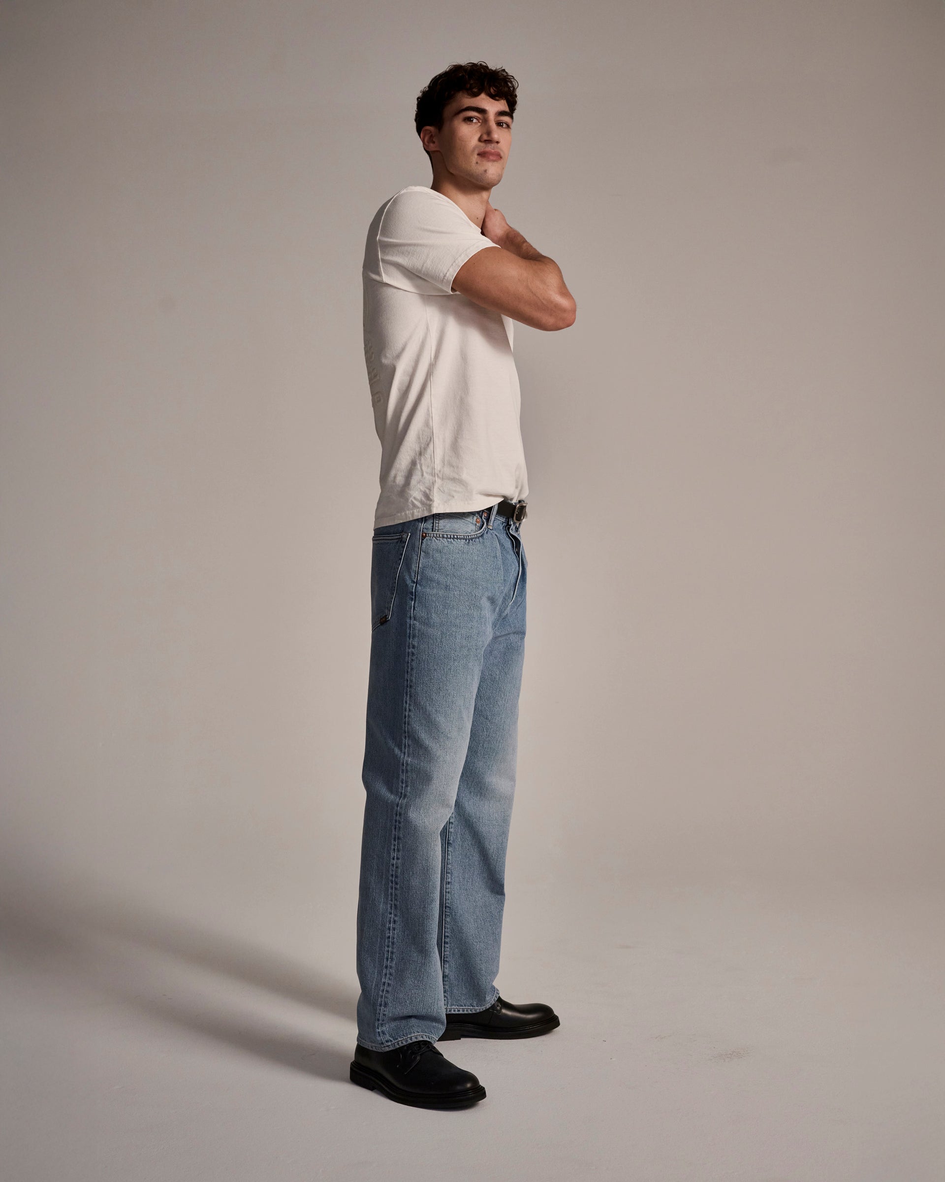 A young man stands sideways in a studio, wearing T7C’s The Cinch Jean in Mykonos with a white t-shirt and black shoes, hand on his neck and gazing upward against a plain background.