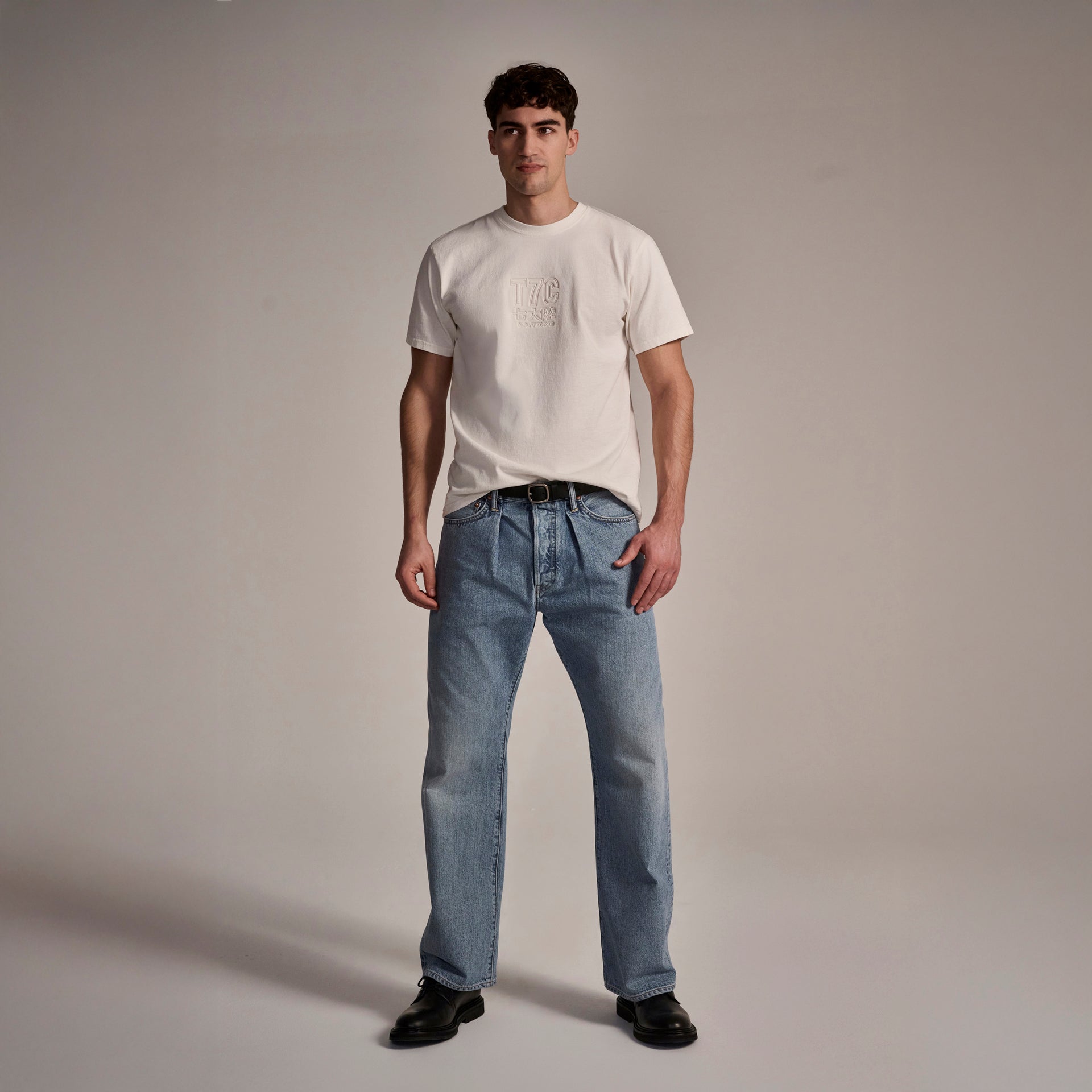 A man stands facing forward in a plain studio, wearing T7Cs The Cinch Jean in Mykonos with a white t-shirt, black belt, and black shoes. The neutral background is uncluttered.