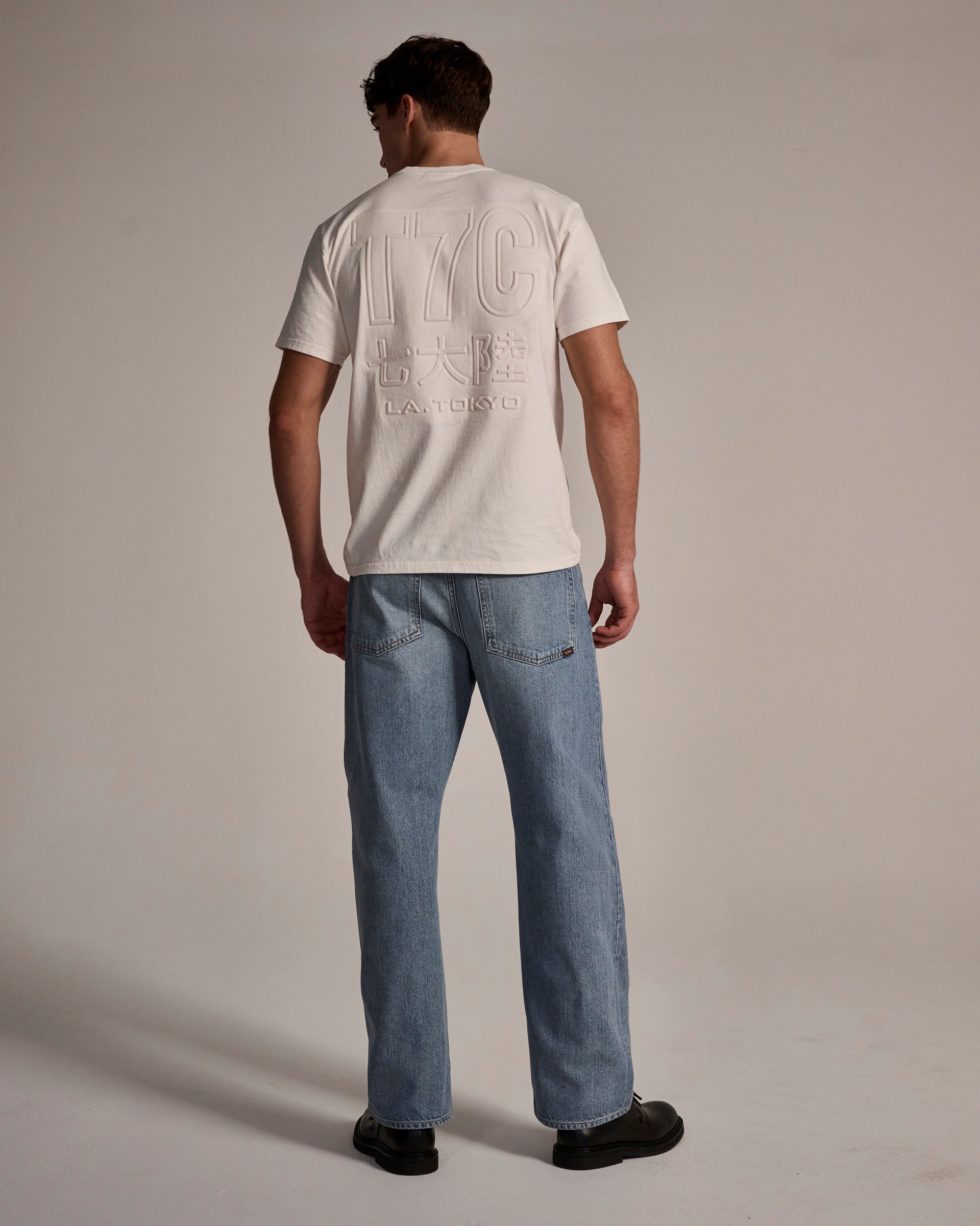 A man faces away against a light background, wearing a white tee with embossed back graphics, T7C The Cinch Jean in Mykonos (light blue selvedge denim), and black shoes.