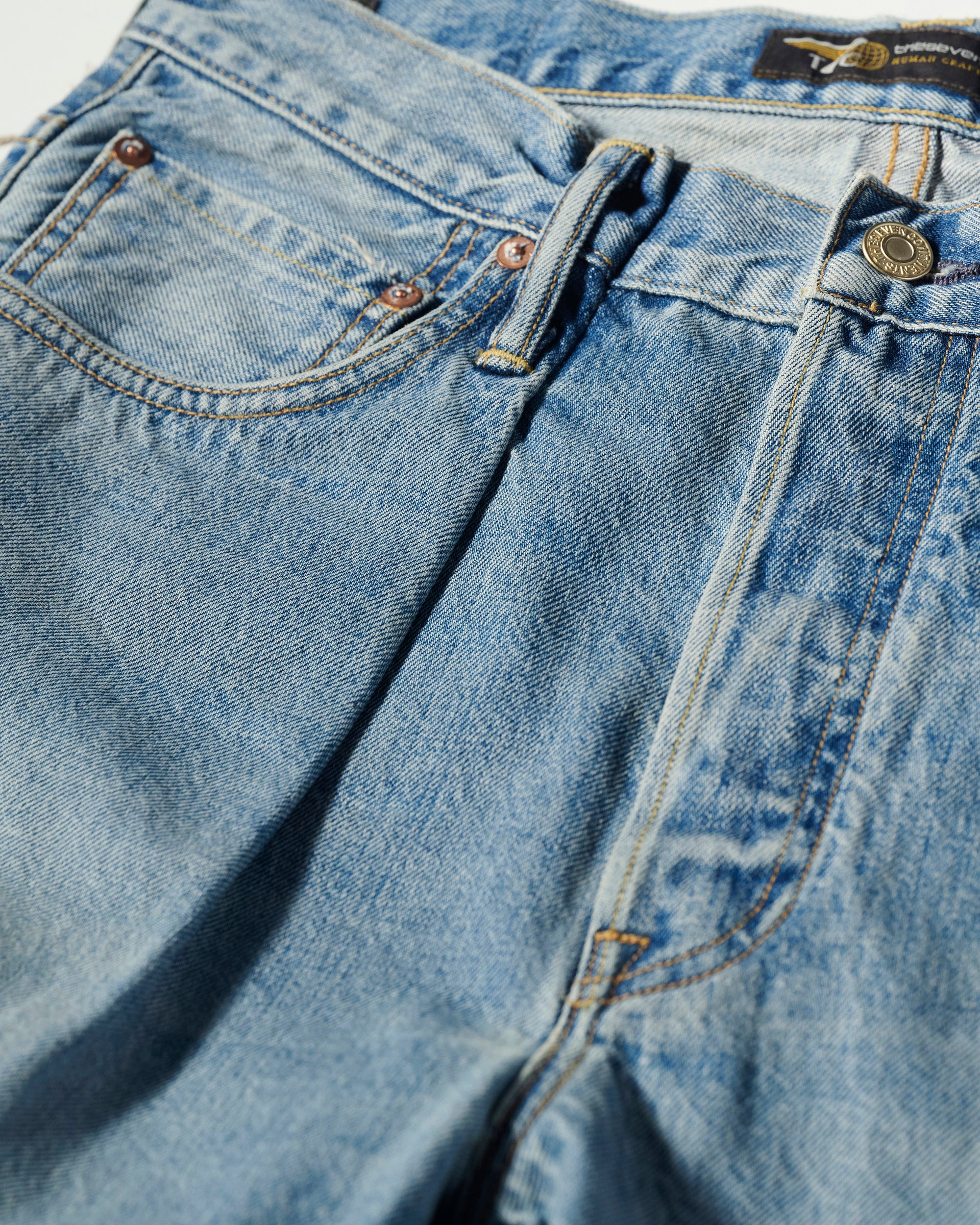 Close-up of T7Cs The Cinch Jean in Mykonos, featuring front pockets, belt loops, zipper, button, signature stitching, and a light blue vintage-worn selvedge denim texture.