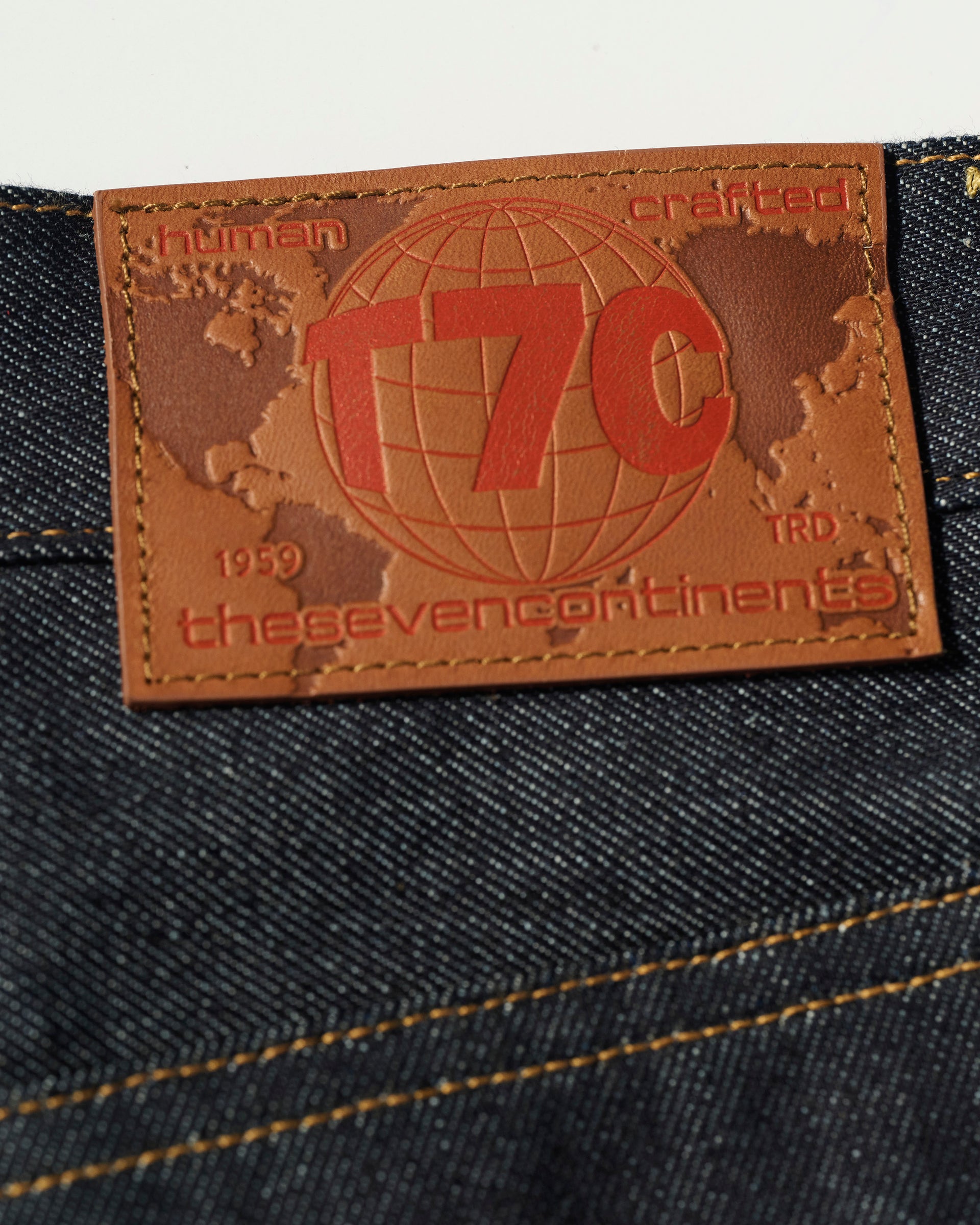 A close-up of The Cinch Jean in Yokohama Raw by T7C The Seven Continents shows a brown leather globe patch with “T7C, human crafted, thesevencontinents, 1959, TRD” and standout orange stitching against rich indigo-dyed denim.