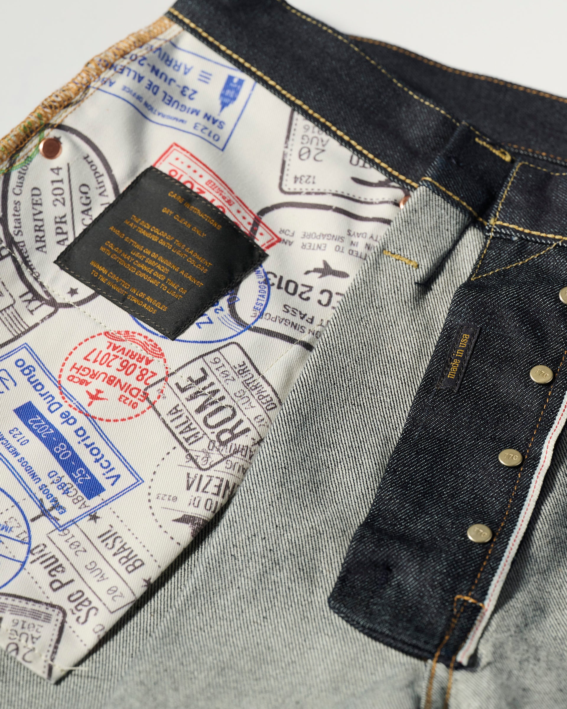 Close-up of The Cinch Jean in Yokohama Raw by T7C The Seven Continents, turned inside out to show a white lining with colorful passport stamps, contrasted by dark indigo denim, visible stitching, metal rivets, and buttons.
