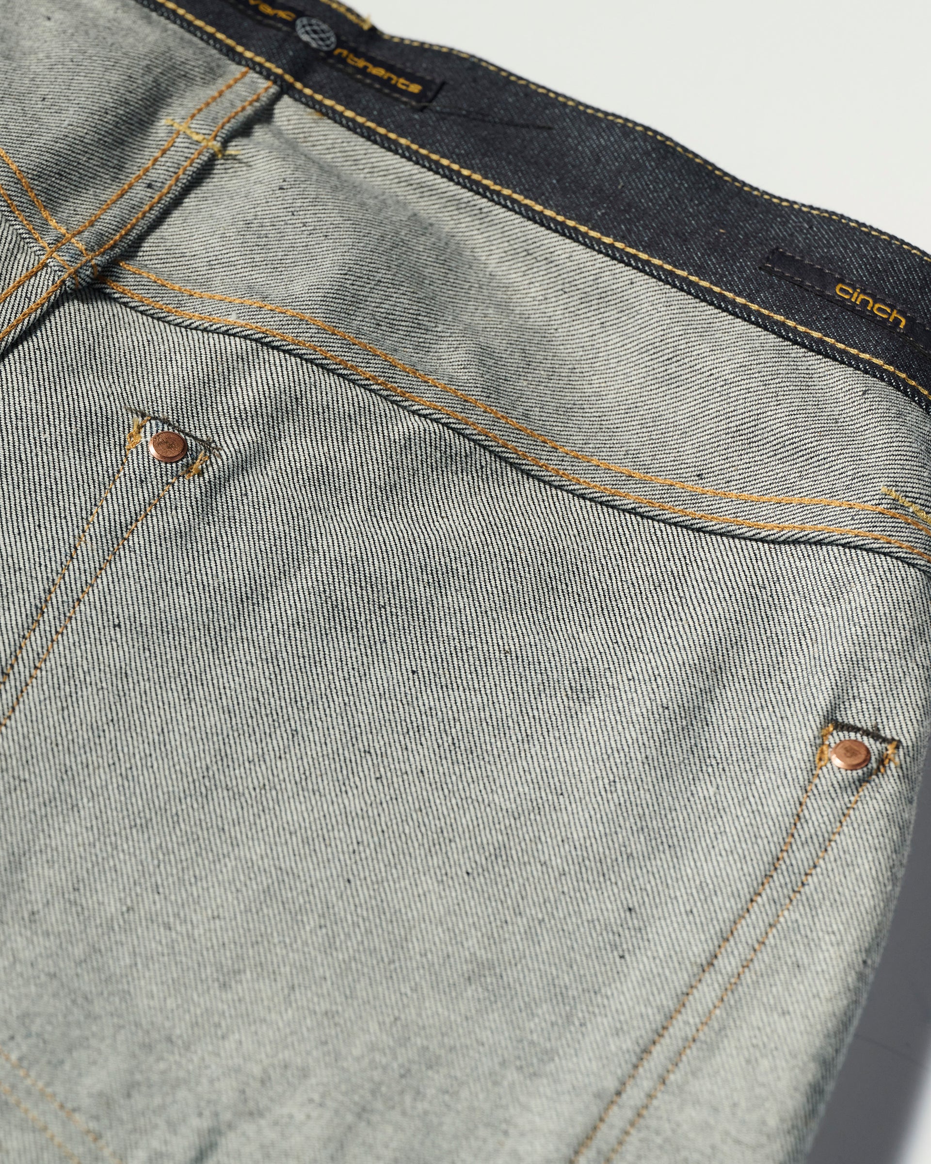 Close-up of The Cinch Jean in Yokohama Raw by T7C The Seven Continents, featuring light blue indigo-dyed denim, orange stitching, copper rivets, and a visible cinch waistband label with detailed fabric texture and pocket accents.