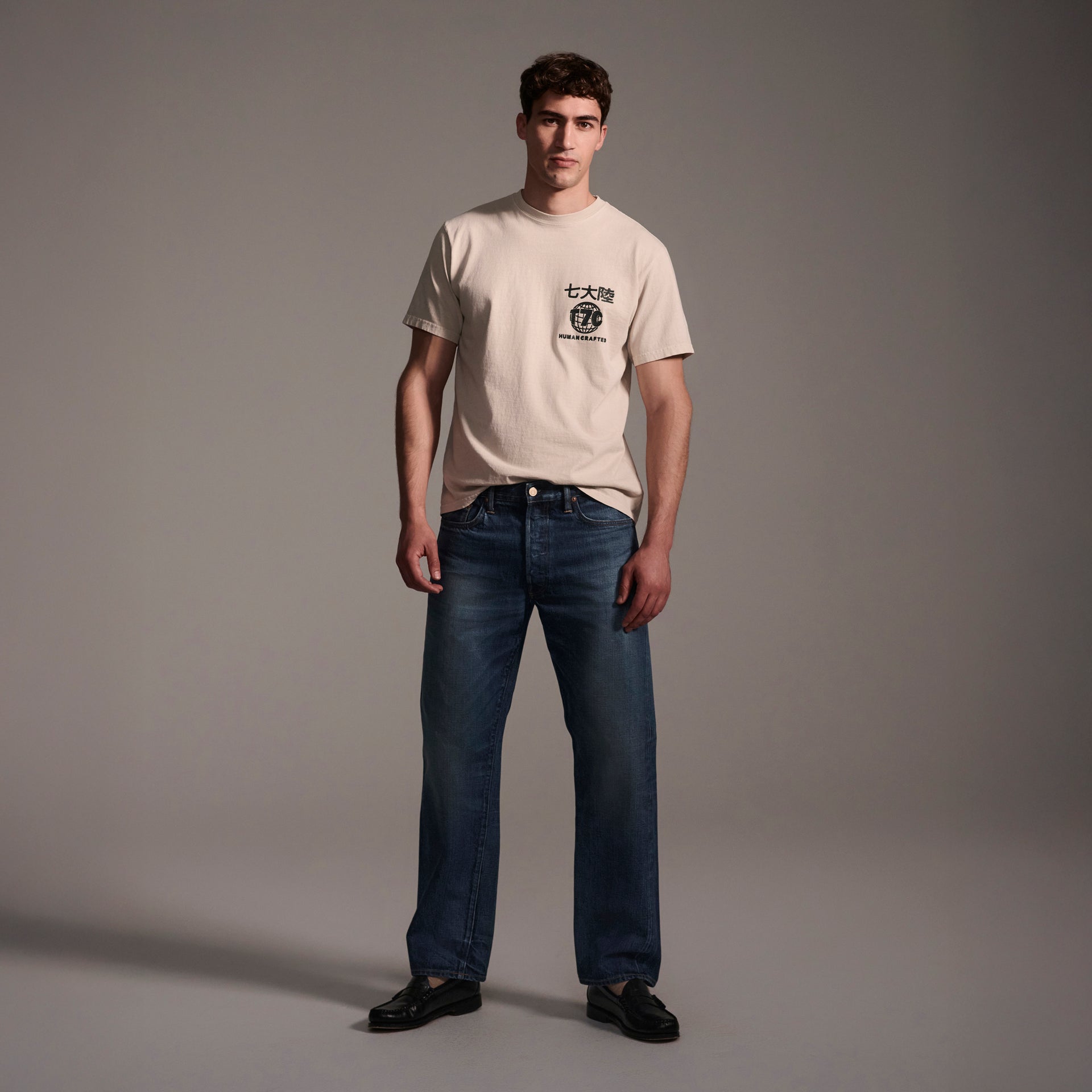A man stands in a studio wearing T7C’s The Dylan Twisted Relaxed Jean in Nimes, a light beige graphic tee with dark text and symbols, and black shoes. The background is plain gray with soft lighting.