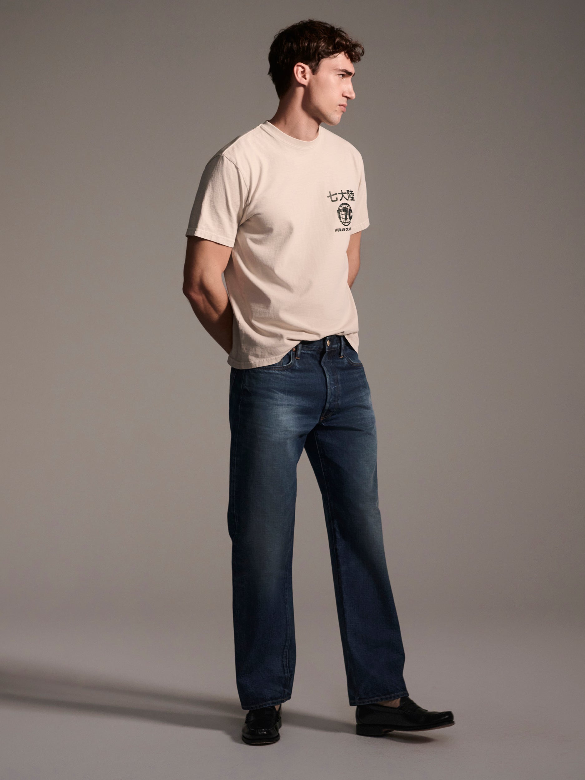 A man stands against a plain gray background, hands behind his back, wearing a light beige graphic tee, black shoes, and T7Cs The Dylan Twisted Relaxed Jean in Nimes.