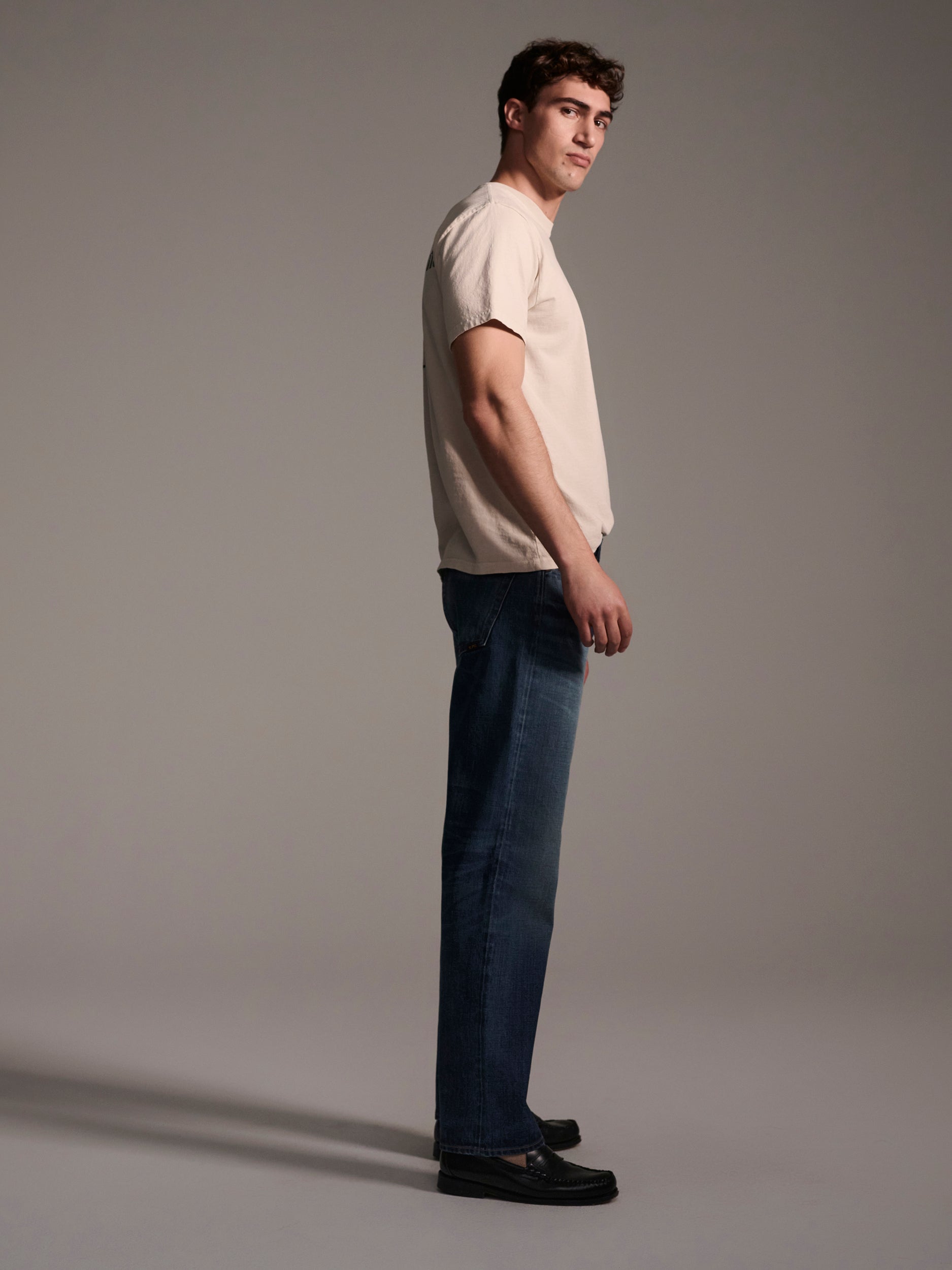 A man stands in profile against a plain background, wearing a light beige t-shirt, T7C’s The Dylan Twisted Relaxed Jean in Nimes, and black shoes.