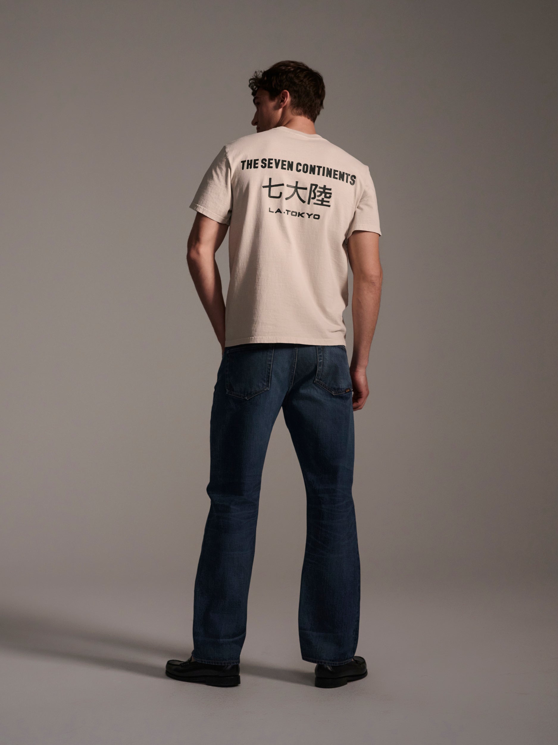 A man stands facing away, wearing a light-colored T-shirt with THE SEVEN CONTINENTS and Japanese text on the back, paired with T7C The Dylan Twisted Relaxed Jean in Nimes and black shoes, set against a plain studio background.