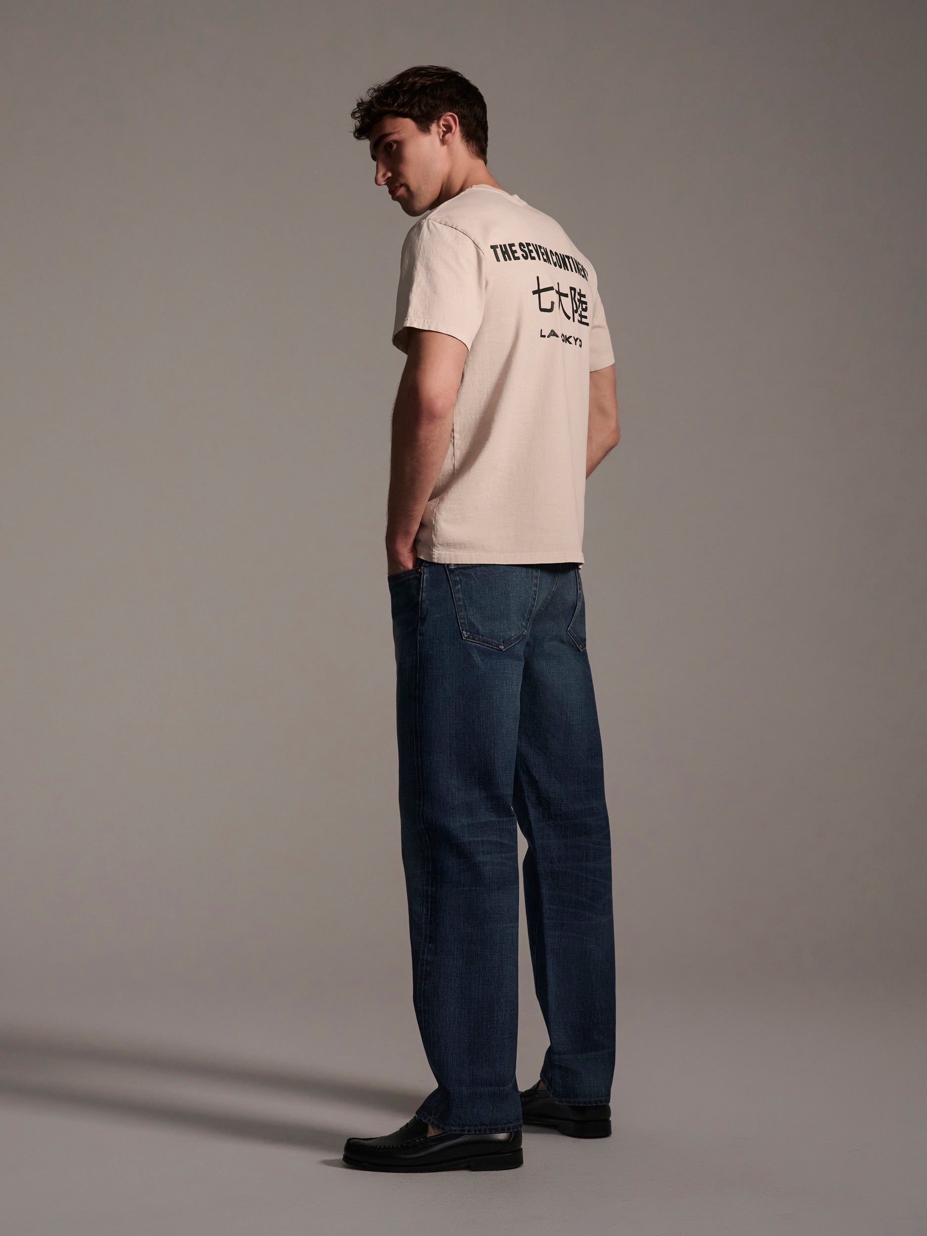 A young man stands sideways, looking over his shoulder. He wears a beige tee and T7C’s The Dylan Twisted Relaxed Jean in Nimes, paired with black shoes, against a plain neutral background.
