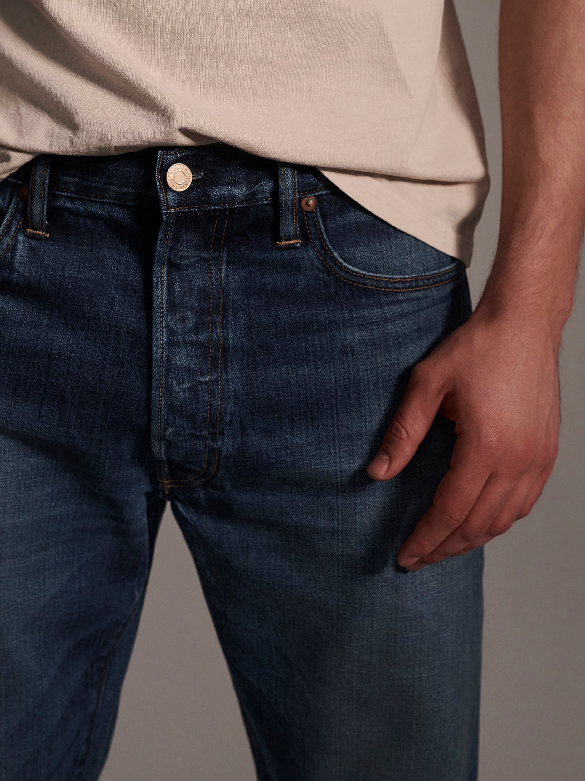 A person wearing a light beige t-shirt and T7Cs The Dylan Twisted Relaxed Jean in Nimes is shown from waist to mid-thigh against a neutral background, with one hand resting by their side.