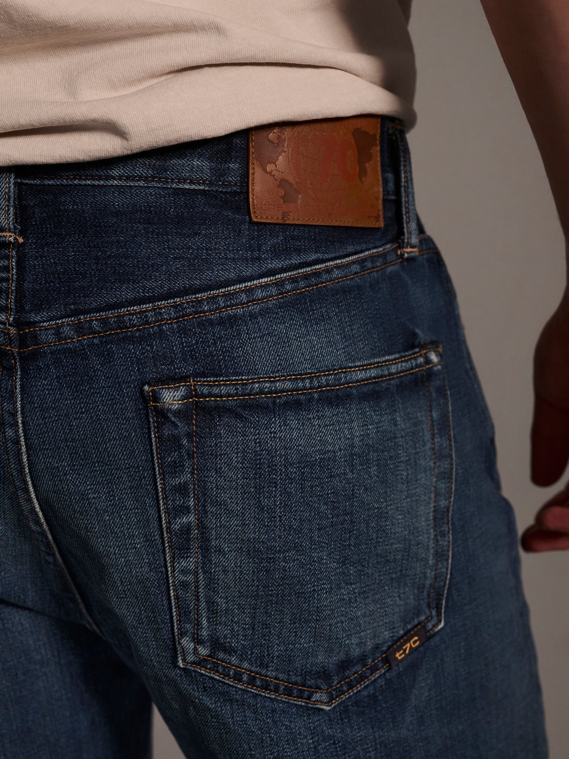 Close-up of a person wearing T7Cs The Dylan Twisted Relaxed Jean in Nimes, paired with a beige shirt. The image highlights the back pocket, leather brand patch on the waistband, and the persons right arm at their side.