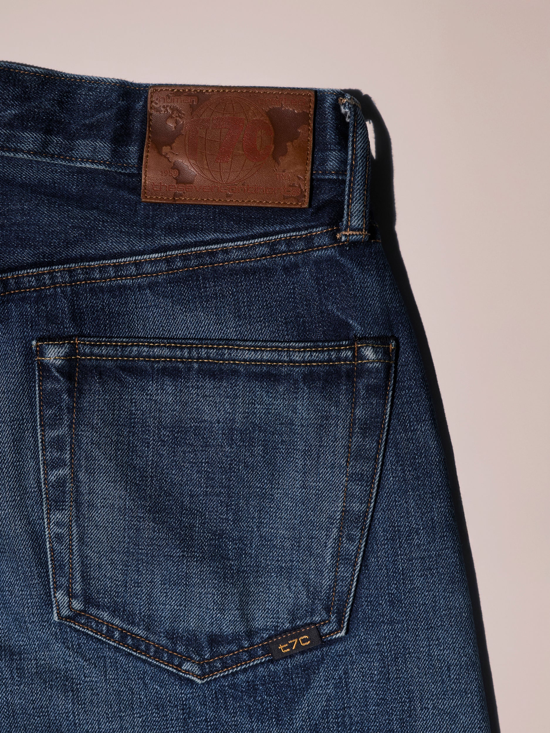 A close-up of the T7C Dylan Twisted Relaxed Jean in Nimes shows the back pocket and waistband with a brown embossed leather patch and visible stitching, set against a light background.
