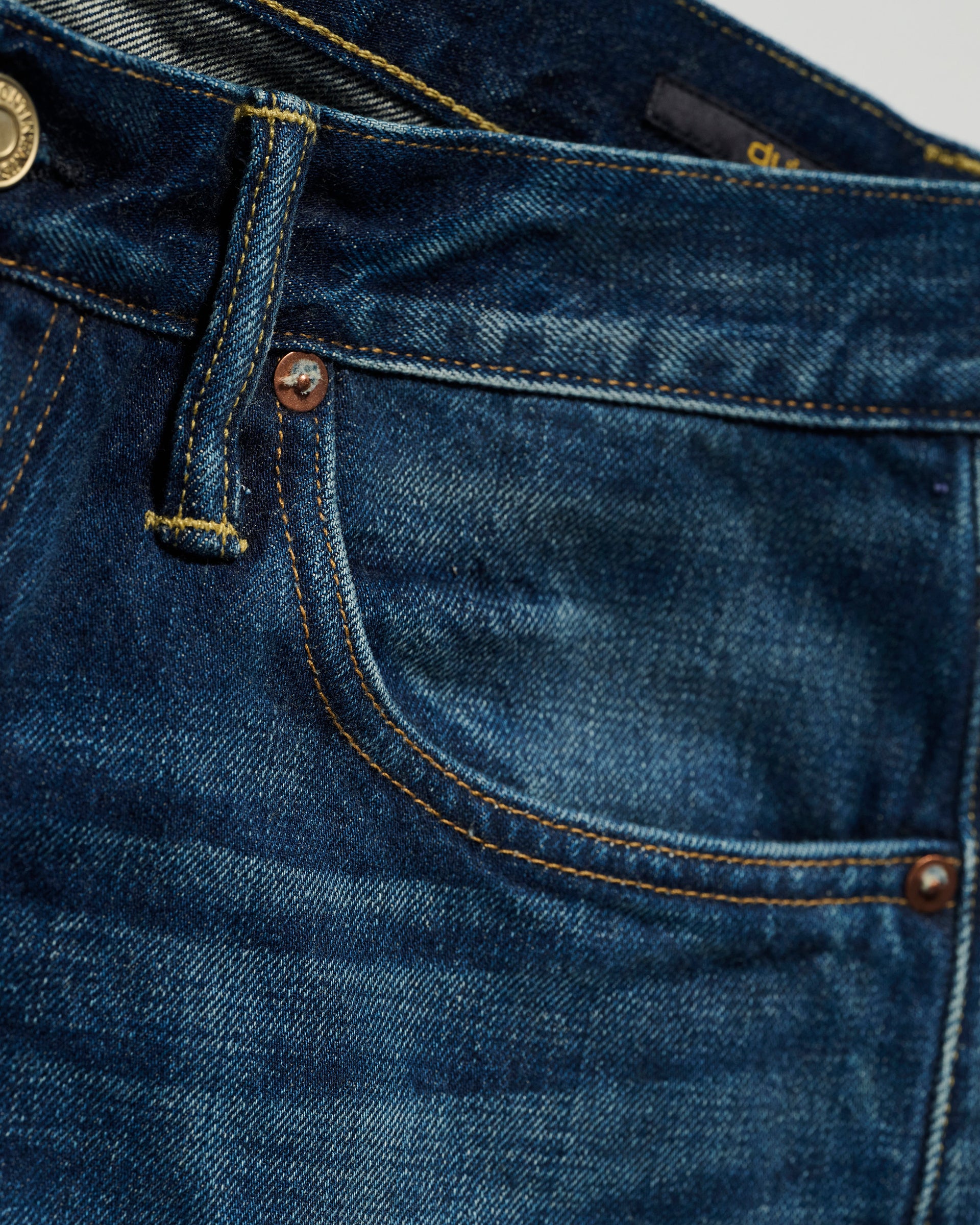 Close-up of the upper section of T7Cs The Dylan Twisted Relaxed Jean in Nimes, highlighting the waistband, front pocket, belt loop, copper rivets, and metal button with visible stitching and a slightly faded denim texture.