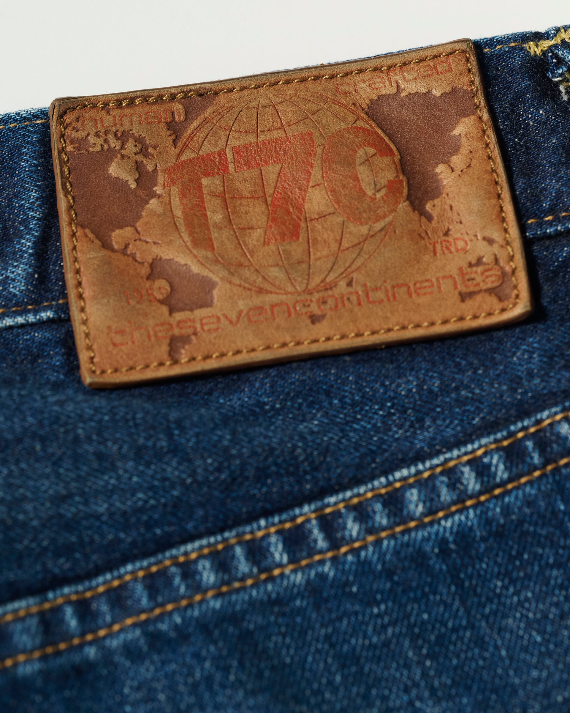 Close-up of a leather patch on The Dylan Twisted Relaxed Jean in Nimes by T7C. The patch displays a world map, T7C, and the seven continents, with yellow stitching on the vintage blue denim fabric around it.