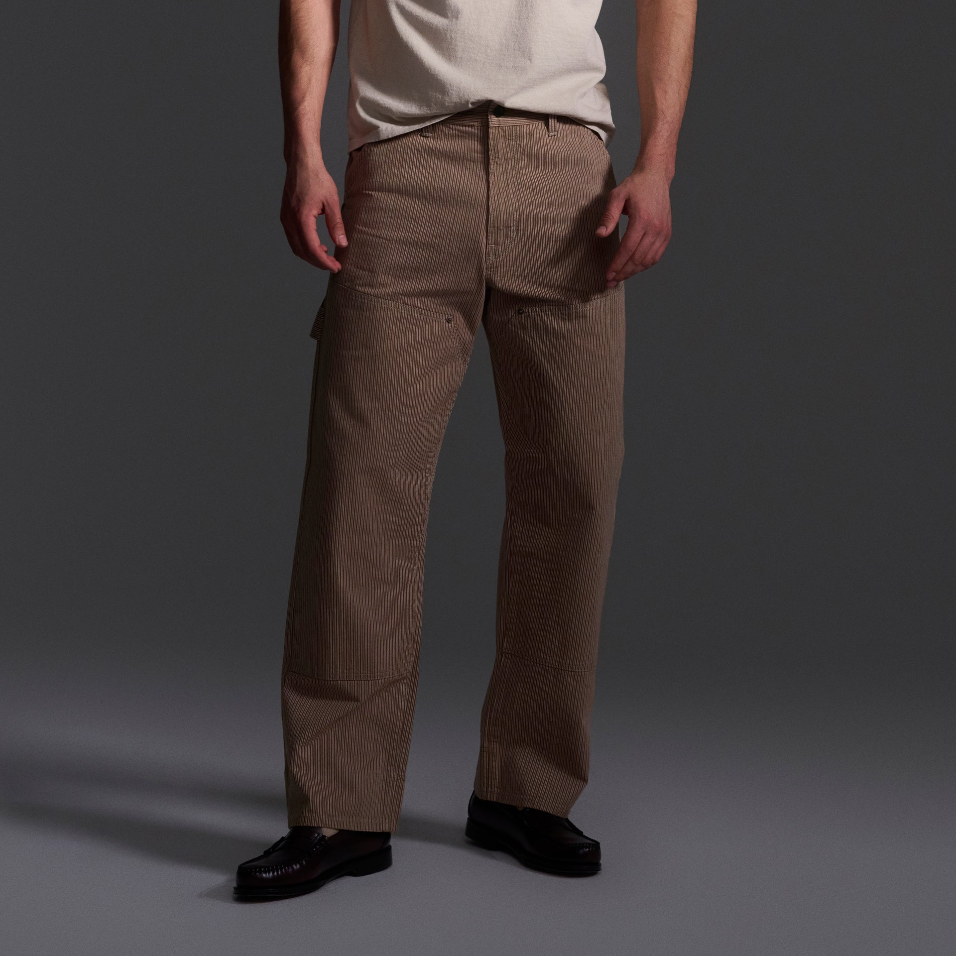Only the lower half of a person is visible, wearing T7Cs The Fred Pant in Camel Stripe—relaxed-fit carpenter pants made from Japanese mill fabric—paired with a light-colored t-shirt and black shoes, set against a dark background.