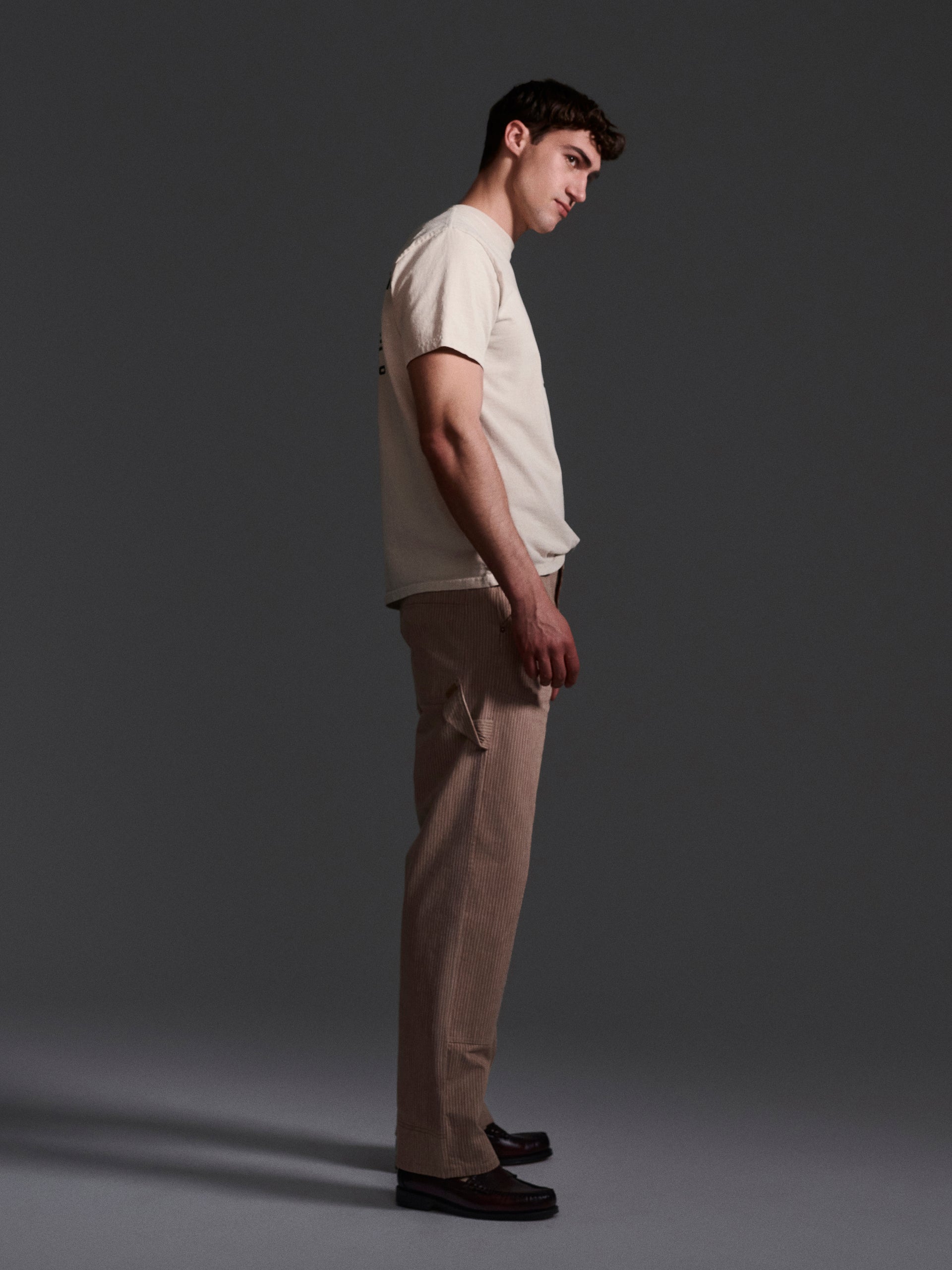 A young man stands in profile against a dark background, wearing a beige T-shirt, T7C’s The Fred Pant in Camel Stripe, and dark loafers. He looks slightly downward with a neutral expression.