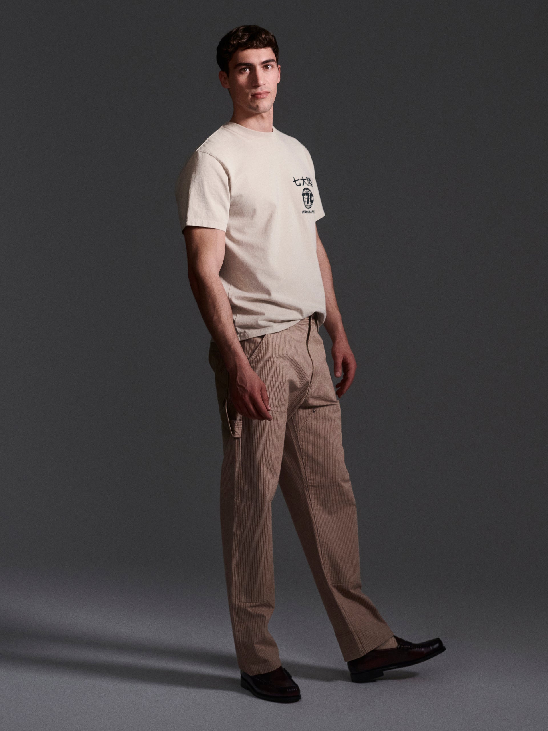 A man stands against a dark background, wearing a beige T-shirt with a small chest graphic, T7Cs The Fred Pant in Camel Stripe made from Japanese mill fabric in a relaxed fit, and black shoes. He faces slightly to the side with a neutral expression.