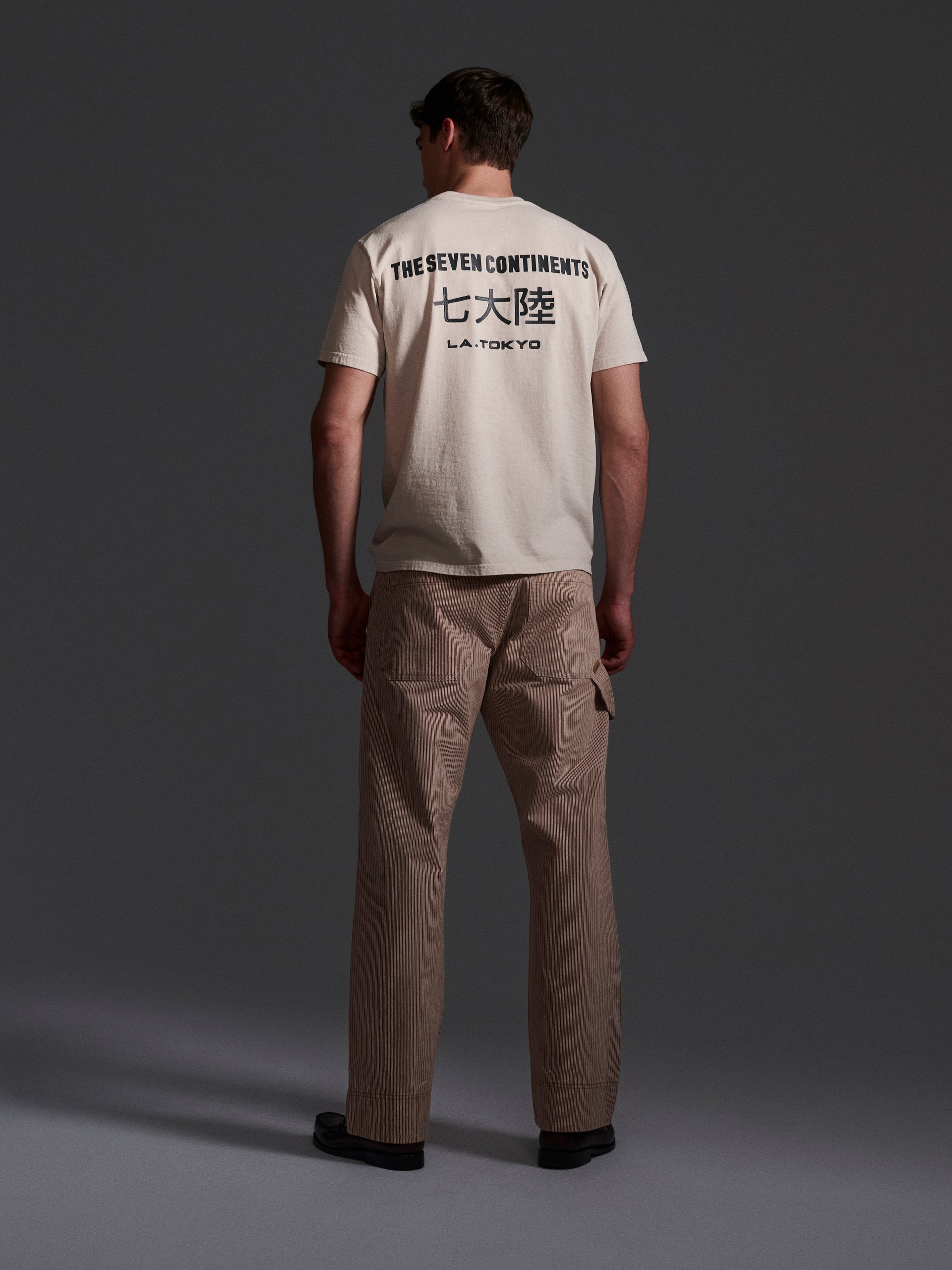 A man faces away, wearing a cream T-shirt with THE SEVEN CONTINENTS and Japanese text above LA TOKYO, paired with T7C’s The Fred Pant in Camel Stripe, relaxed-fit carpenter pants made from premium hickory railroad stripe fabric.
