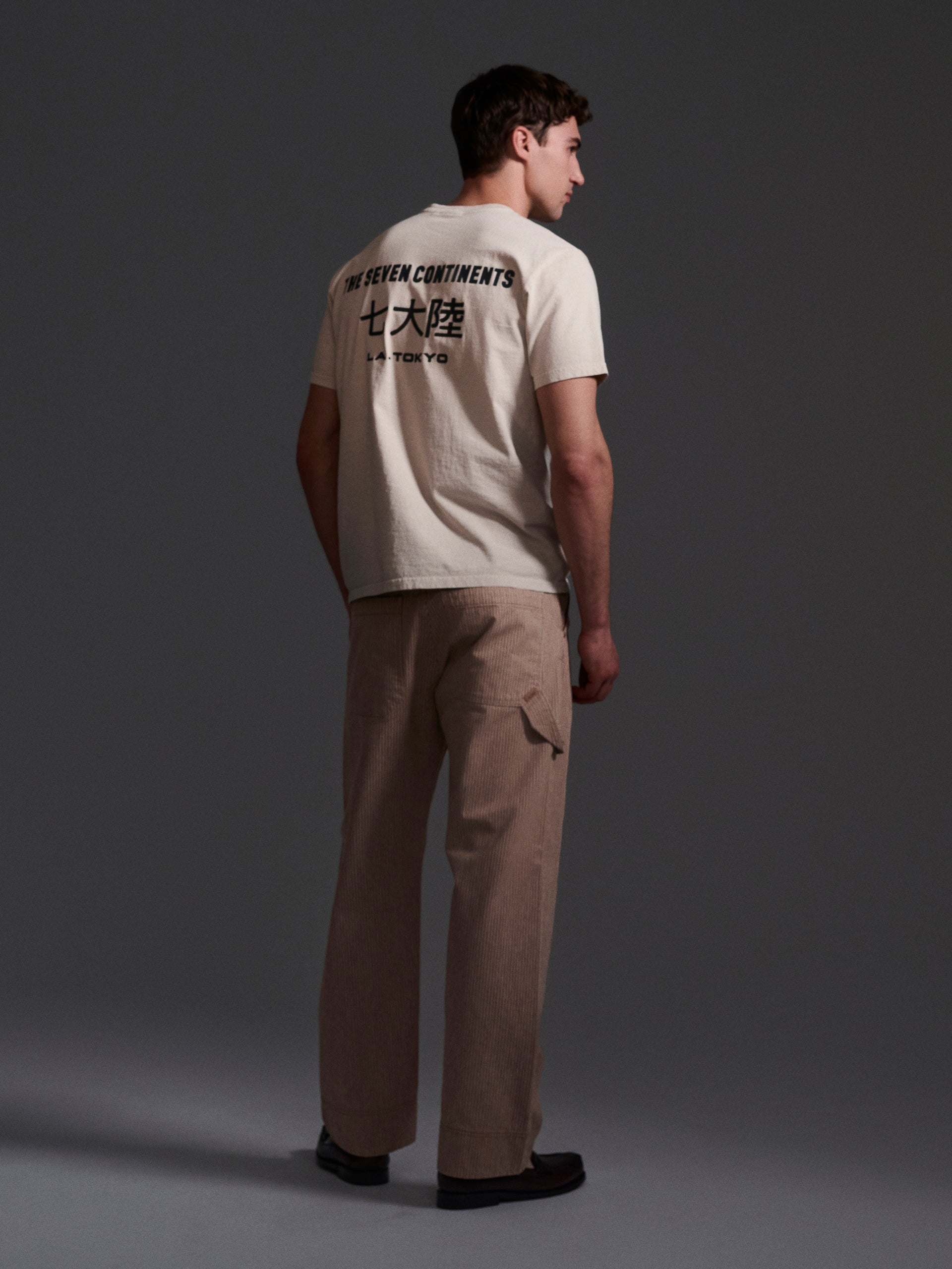 Facing away, a man wears a beige T-shirt with text and Chinese characters on the back, paired with T7C’s The Fred Pant in Camel Stripe—relaxed-fit carpenter pants from a Japanese mill—and dark shoes, against a plain dark backdrop.