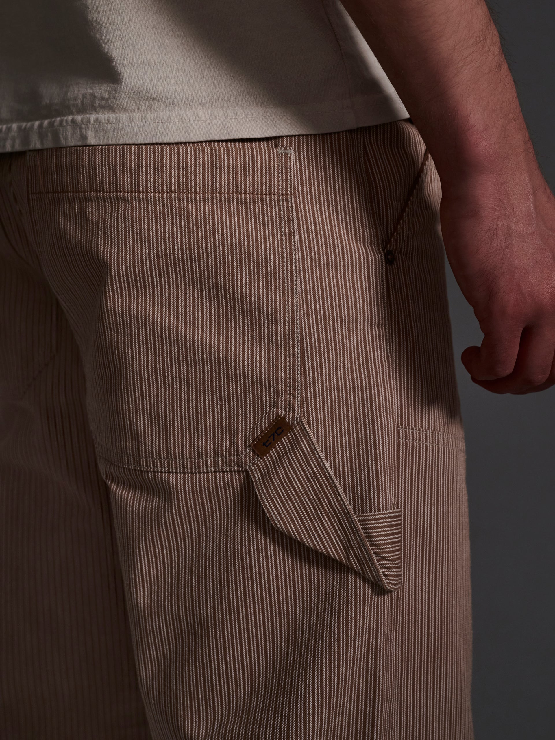 A close-up shows a person in T7Cs The Fred Pant in Camel Stripe—relaxed-fit carpenter pants with beige and white classic hickory railroad pinstripes. They stand sideways, arm at their side, wearing a light shirt against a dark background.