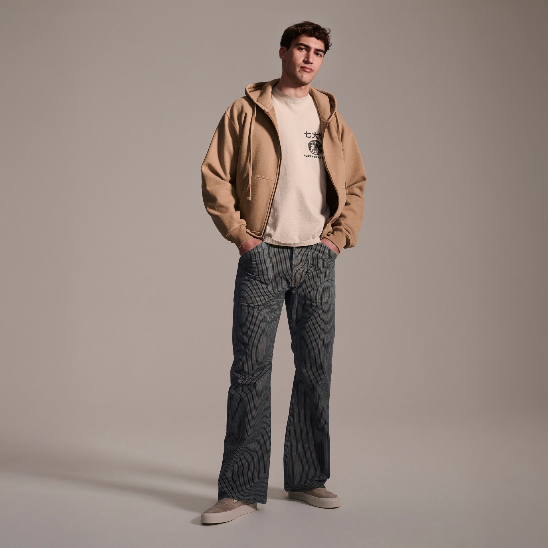 A young man stands confidently against a plain background, wearing a tan zip-up hoodie over a light T-shirt, and T7C The Seven Continents Grayson Pant in Indigo Stripe, with his hands in his pockets as he faces the camera.